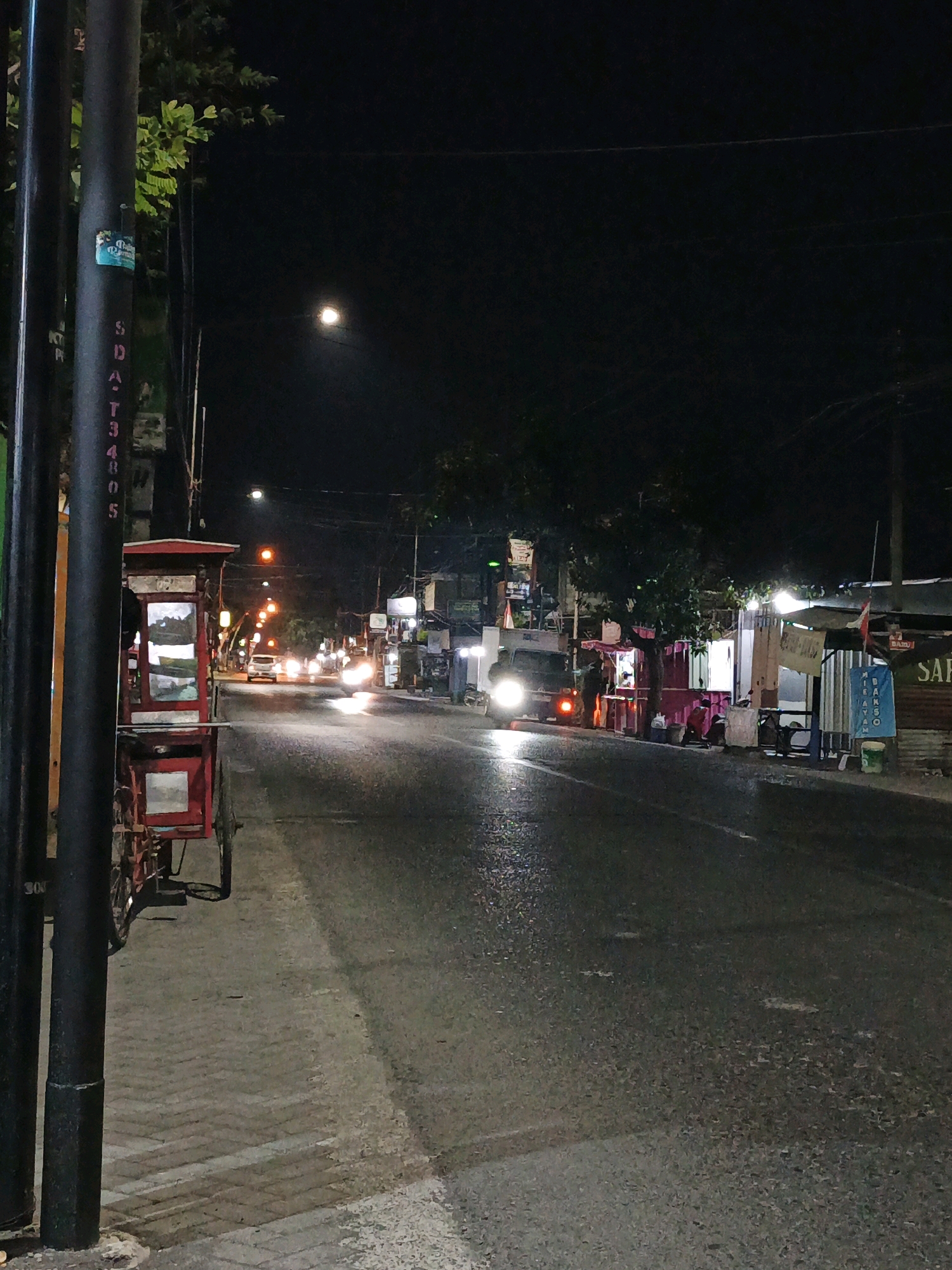 This is a night-time photograph of a street scene in Randegan, Indonesia. The image is dominated by a dark sky with a faint illumination from a distant light source, possibly the moon or a streetlamp, visible in the upper center. A paved road occupies the majority of the frame, wet and reflective, indicating recent rain or high humidity. Traffic lights emit orange and white glows along the road, and the headlights of vehicles create streaks of light.
On the left side of the road, a dark pole stands prominently, with some green foliage visible behind it. Attached to the pole are stickers with what appears to be text. A red and white cart, possibly a food stall or vendor's cart, is parked alongside the sidewalk, which is constructed of interlocking pavers.
The right side of the road features a row of small buildings or stalls, some with open fronts and illuminated interiors. Figures of people can be seen near these establishments, suggesting they are open for business. Signs are visible, including one that appears to say "SANT," and another with blue text that may be a menu or advertisement for "MIE AYAM." The overall atmosphere is that of a typical bustling street in a populated area at night, with ongoing commerce and activity despite the darkness.