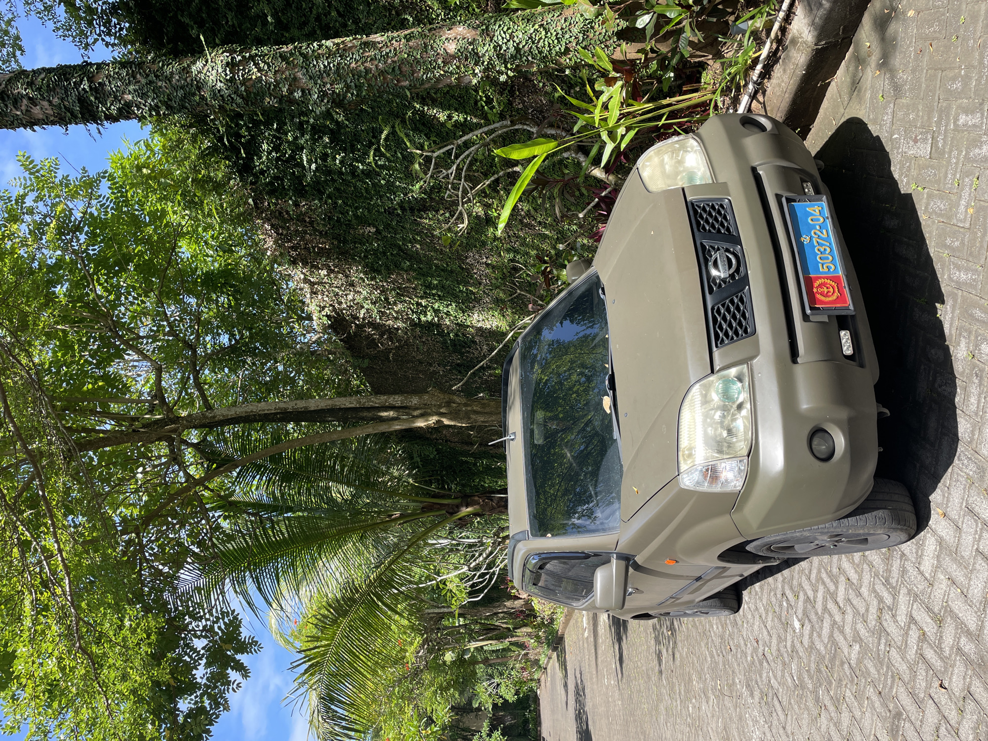 A khaki-colored Nissan X-Trail SUV is parked on a brick road next to a lush, green tropical environment. The vehicle's license plate reads "50372-04" and features a red and blue color scheme with a coat of arms. The car is likely in Kuta Selatan, Indonesia, based on the tropical foliage. Trees with dense vines climb up their trunks, and palm trees are visible. The sky is blue with scattered clouds. The overall setting appears to be a residential area or a park in a tropical climate.