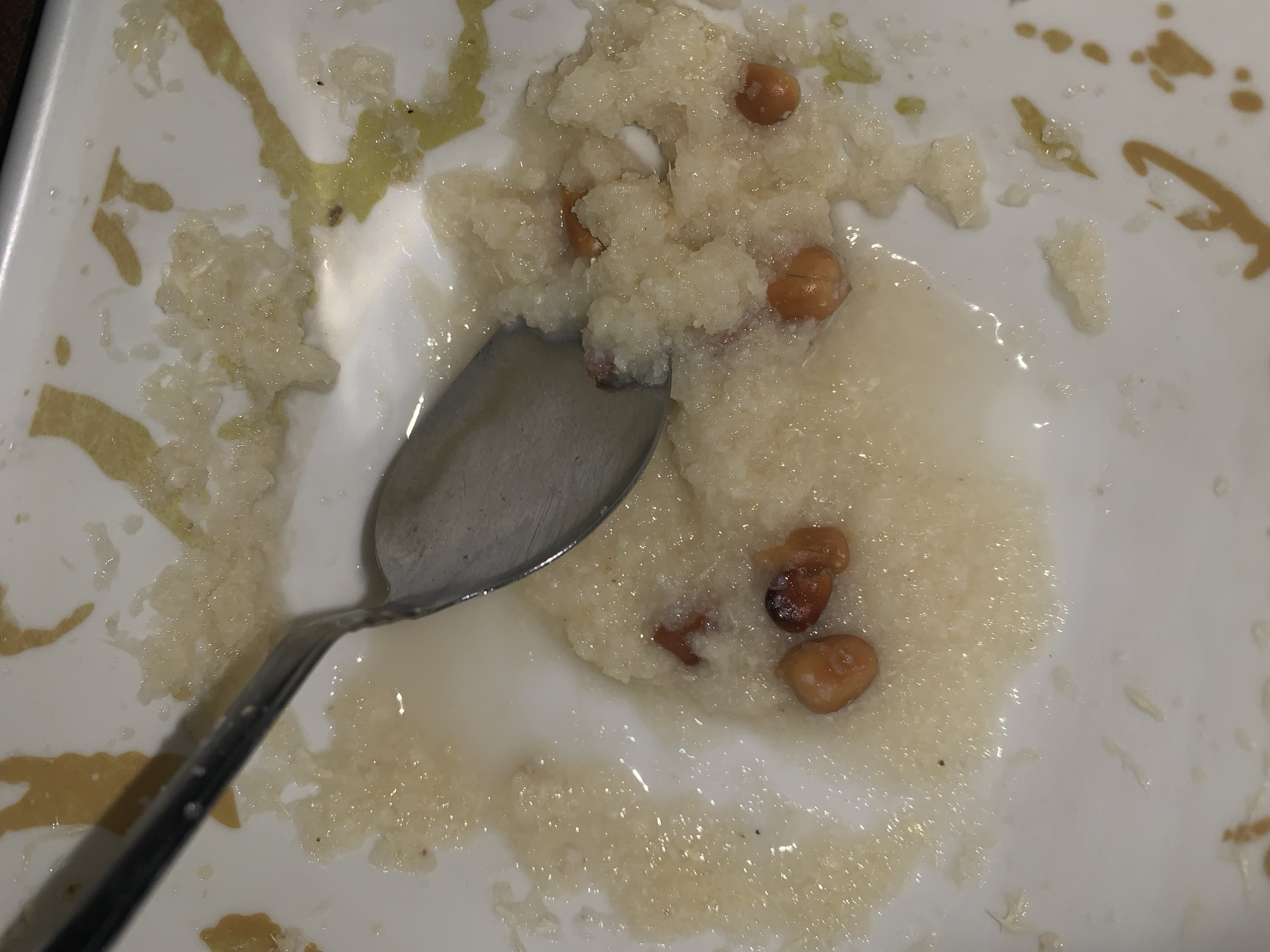The image provides a close-up, top-down view of a partially consumed meal served on a white plate, with a silver spoon resting within the food. The primary subject is a light-colored, granular, semi-liquid dish, resembling a porridge or gruel. This main food item is interspersed with several reddish-brown, oval-shaped pieces, which are consistent with roasted peanuts, adding both texture and flavor to the dish.
The characteristics of the food, particularly its porridge-like consistency and the inclusion of peanuts, suggest it could be a traditional Nigerian dish such as 'pap' (akamu or ogi) or 'kunu', which are common cereal-based porridges, often enjoyed for breakfast or as a light meal. Peanuts are frequently used in Nigerian cuisine, either as an ingredient or a garnish.
The white ceramic plate is not pristine; it exhibits distinct greenish-yellow and brownish-yellow streaks and smears around the edges and in areas not covered by the main food item. These residues might be from a sauce, a condiment, or perhaps remnants of another dish previously served on the same plate, indicating an ongoing meal or a less formal dining context.
A metallic spoon is visible, partially submerged in the food on the left side of the plate, confirming that the dish is either being consumed or has just been used. The overall scene suggests a casual dining environment, focusing intimately on the food item. While no explicit visual markers within the image point to Rafin Par, Nigeria, the type of food depicted is culturally consistent with culinary practices in the region. There is no visible text in the media file.