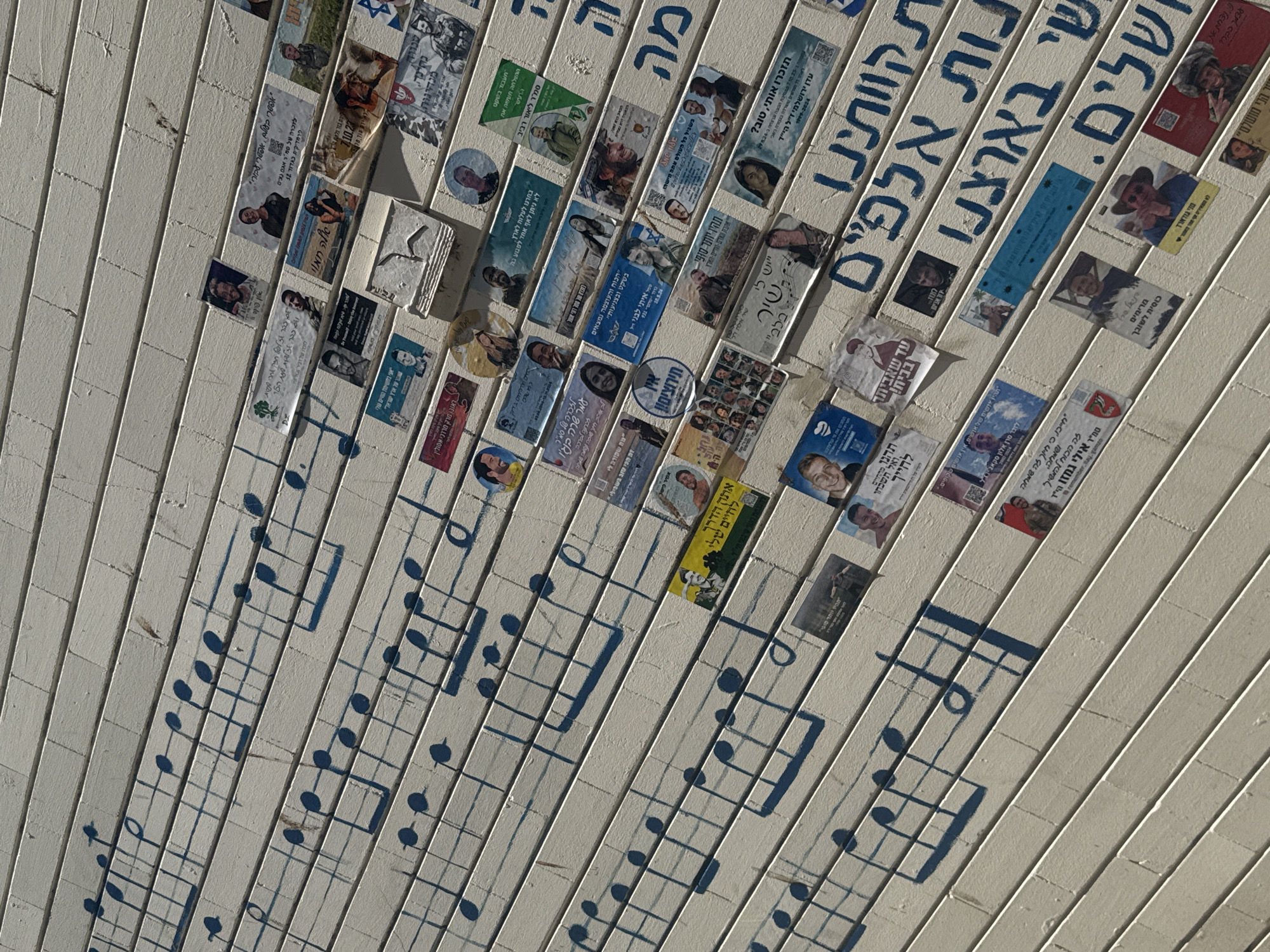 The image depicts a wall or ceiling made of narrow, horizontal, white planks. Affixed to these planks are numerous small photographs and memorial stickers, many featuring faces and Hebrew text. Interspersed amongst these are stylized dark blue musical notes and staves, seemingly painted onto the planks. Text in Hebrew in dark blue is also present and reads "תות אלפיים," "ארצנו," and "שלים." The overall impression is of a memorial or tribute wall in a public space, potentially located in Tel-Aviv, Israel.