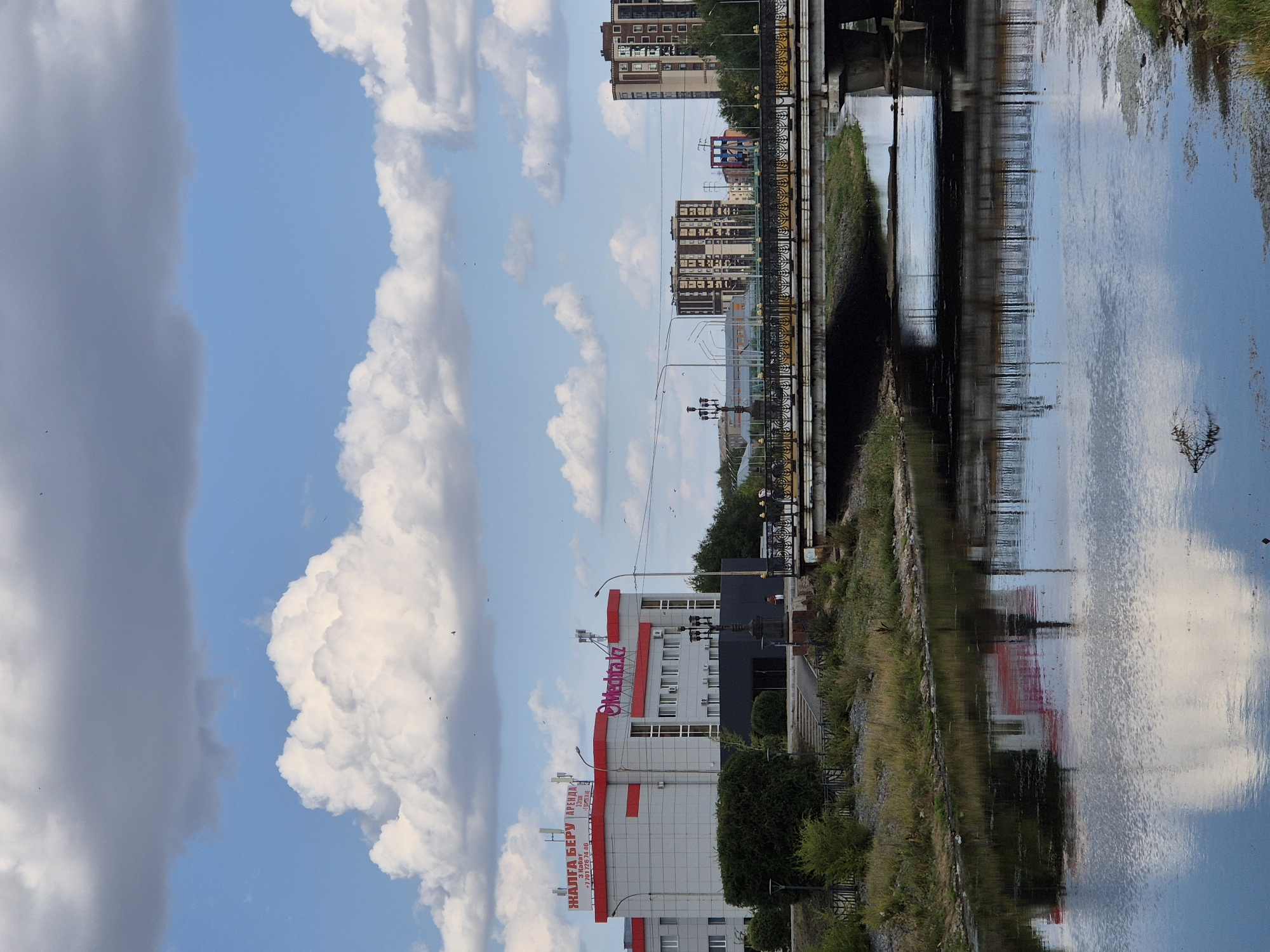 The image shows a river flowing through the city of Kokshetau, Kazakhstan. A bridge spans the river, with a modern building on the left bank and several apartment buildings on the right bank. The building on the left has signage in Kazakh, indicating it is a commercial establishment. The river's surface reflects the sky and the bridge. The sky is partly cloudy with large, white cumulus clouds. The overall scene suggests an urban environment during the daytime.
