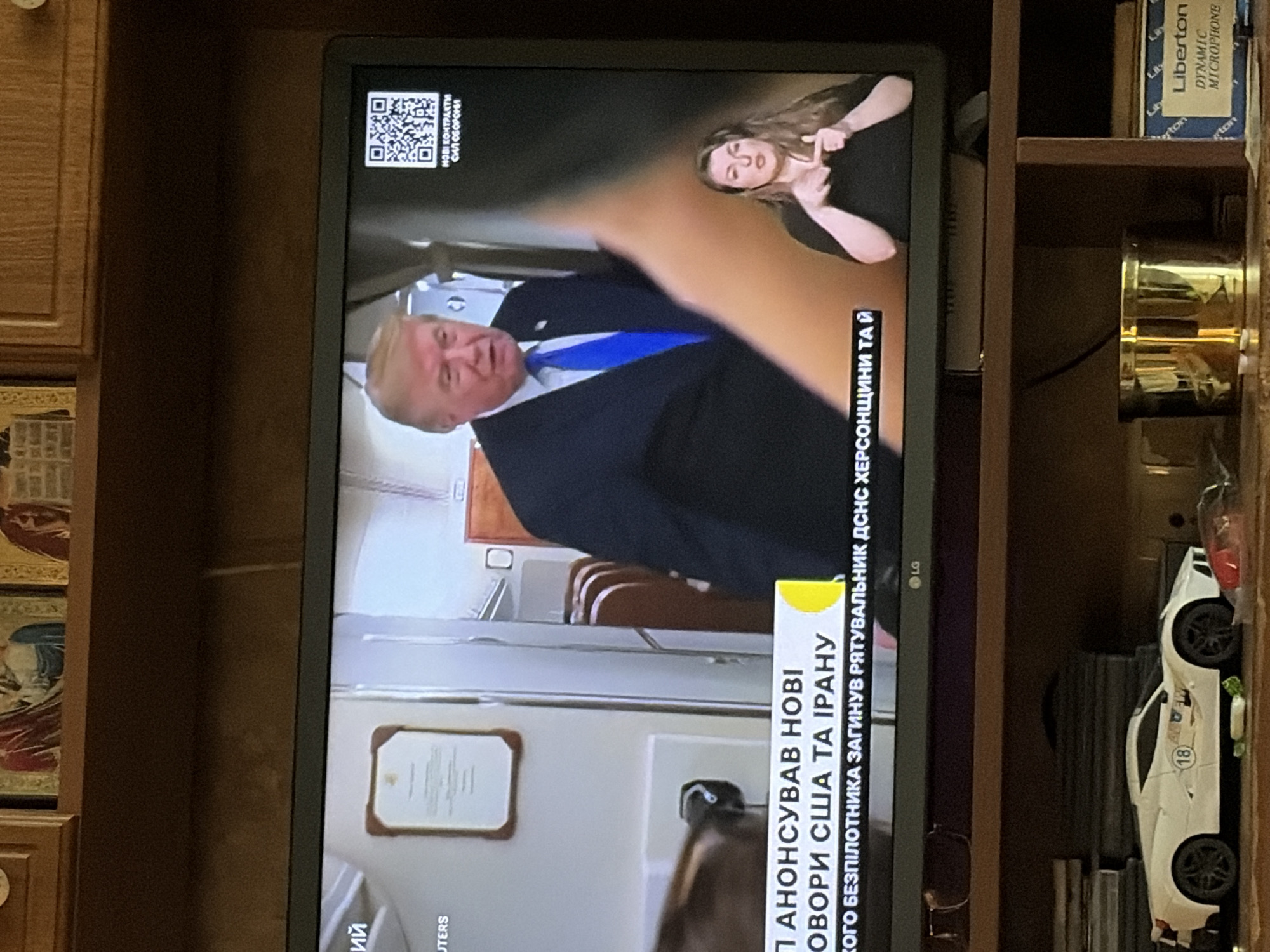A television displays a news report featuring Donald Trump in a suit and tie. The chyron text below him reads "АНОНСУВАВ НОВІ...". To the right, a woman is signing on screen. In the lower portion of the screen, a white toy car is visible on a shelf. Above the television, religious icons are displayed on a wooden cabinet. The scene appears to be an indoor setting.