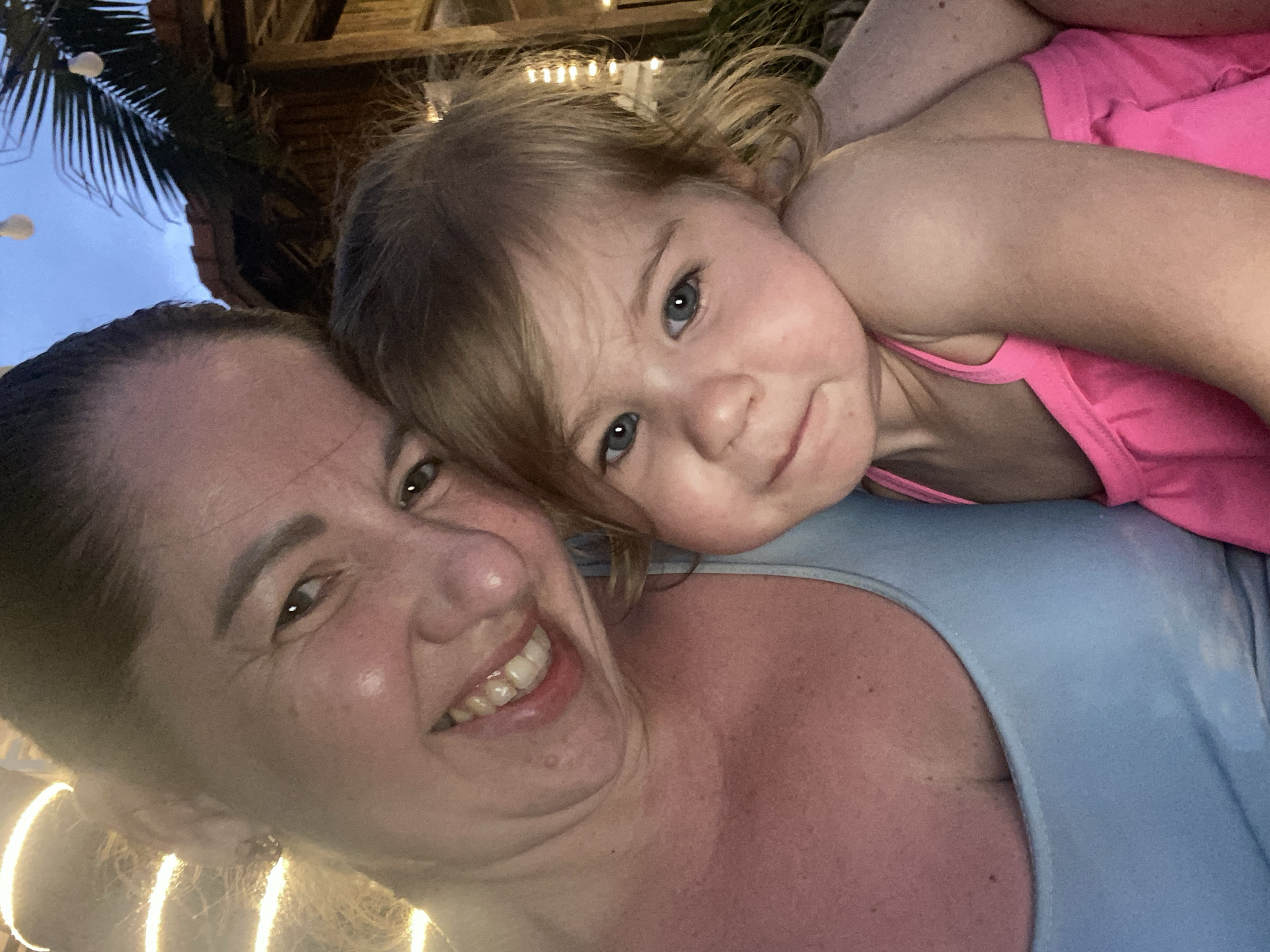 This close-up, slightly tilted selfie captures an adult woman and a young child in an outdoor setting at dusk or early evening. The woman, positioned in the foreground and to the left, smiles broadly at the camera, showing a cheerful expression with visible smile lines. She has light brown hair pulled back and appears to be wearing a light blue top. A young child, likely a girl, rests her head affectionately on the woman's shoulder/chest on the right side of the frame. The child has bright blue eyes, light brown, slightly wavy hair, and a playful or content expression, also looking towards the camera. She is wearing a vibrant pink top.
The background reveals an outdoor environment, possibly in Phu Thuy Ward, Vietnam. A dark, twilight sky is visible in the upper left, beneath which a dark palm tree frond is silhouetted. Decorative string lights glow softly in the distance and around the subjects, casting a warm light. Wooden beams or parts of an overhead structure are also discernible in the upper background. The overall mood of the image is affectionate and joyful, capturing a warm moment between the two individuals in a pleasant, illuminated evening atmosphere. There is no visible text in the image.