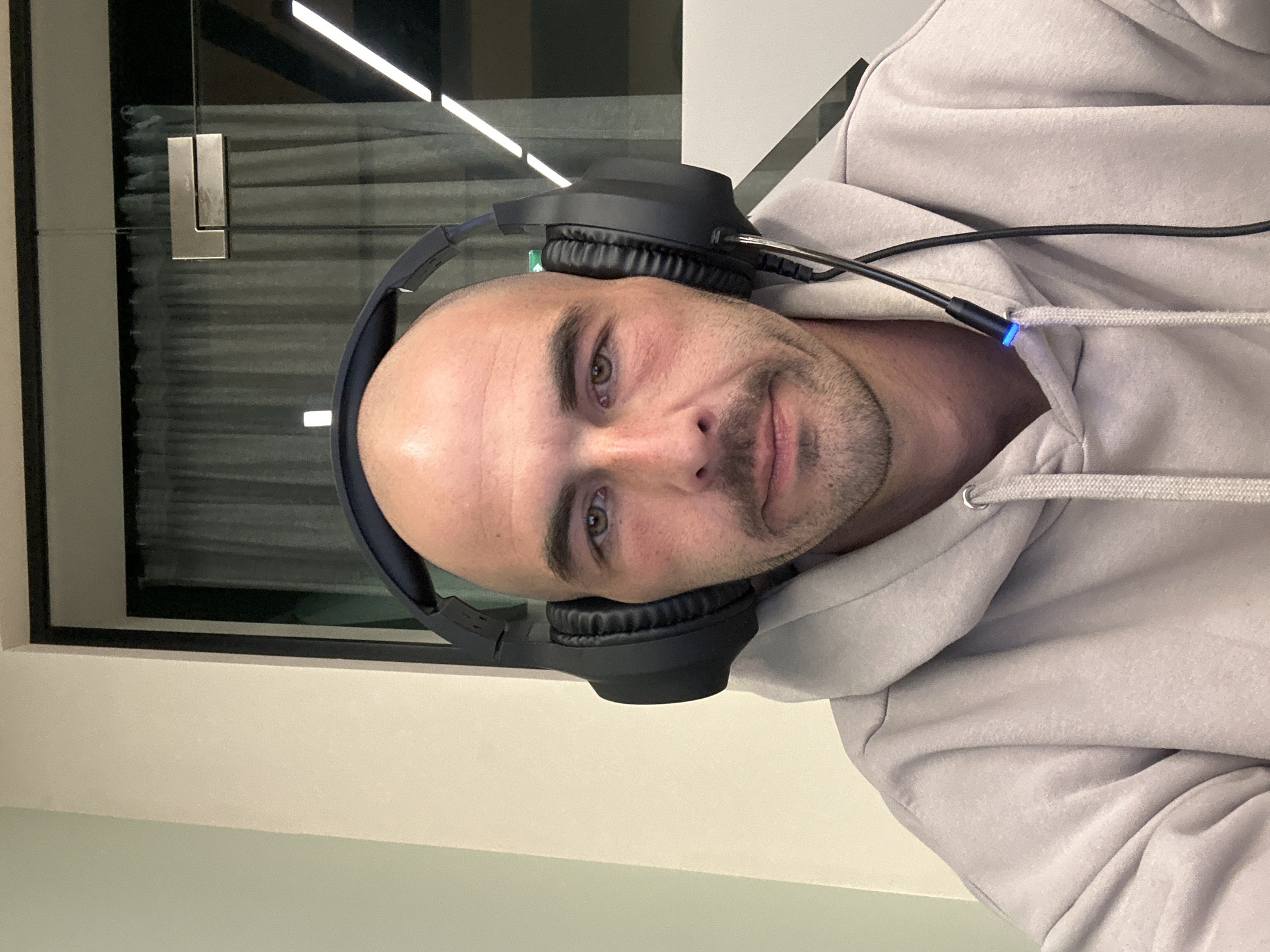 An adult male is depicted in a head-and-shoulders shot, looking directly at the camera in an indoor setting within the city of Budva, Montenegro. The subject is bald, with light skin, brown eyes, a trimmed moustache, and stubble. He is wearing black over-ear headphones, from which a microphone boom extends downwards, featuring a blue illuminated indicator light on its cable near his chest. He is dressed in a light grey hooded sweatshirt. The background consists of a plain light-colored wall on the left and a glass partition on the right. Through the glass, light-colored curtains are visible, along with several horizontal illuminated light fixtures. A metallic, L-shaped fixture is mounted on the glass partition.