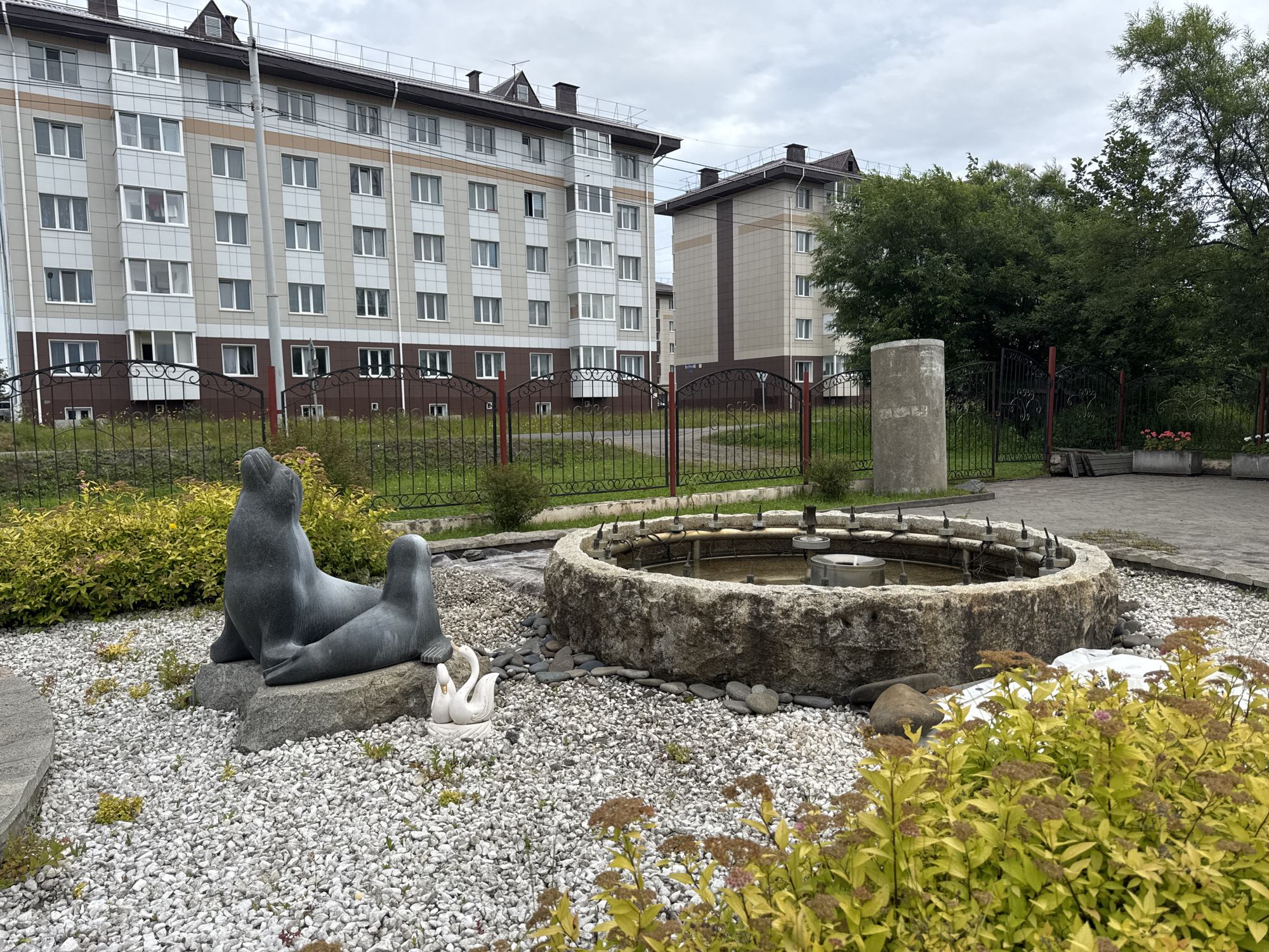 In Poronaysk, Russia, a stone fountain with multiple spray nozzles is situated in a courtyard area. Flanking the fountain, on the left, are two stone sculptures of seals, one larger than the other, positioned on a rocky base. To the right of the seals and in front of the fountain, a small white sculpture of two swans facing each other is visible. The ground around the fountain and sculptures is covered in light-colored gravel, interspersed with small green plants and larger rocks. In the foreground, yellow-leaved bushes with dried brown flower clusters are prominent. Behind the fountain and sculptures, a black metal fence with ornate scrollwork separates the courtyard from a grassy area. Beyond the fence, two modern, multi-story apartment buildings with beige and white facades and dark roofs are visible. The sky is overcast and cloudy. A concrete pillar stands to the right of the fence and behind the fountain.