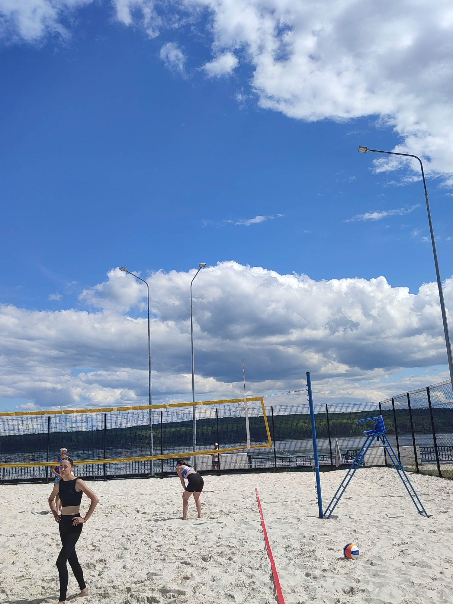 The image depicts a beach volleyball court located in Unknown, Unknown, under a partly cloudy blue sky. The court features fine white sand, a yellow and black mesh volleyball net supported by blue posts, and a red tape line marking a boundary on the sand in the foreground. A multi-colored (white, orange, blue) Mikasa volleyball rests on the sand near this line. To the right of the net, a blue metal referee stand with a seat is visible.
Three individuals are present on the court. In the left foreground, a female with dark hair, wearing a black sports bra and black leggings, stands with her hands on her hips, facing slightly left. In the mid-ground to the right, another female with dark hair, wearing a white sports top and black shorts, is bent over, looking down at the sand. A third individual, wearing a blue top, is partially visible behind the net on the left.
The background shows a tall metal fence with mesh panels separating the court from a large body of water, possibly a river or lake, with a distant treeline on the opposite bank. Three tall metal streetlights with rectangular LED fixtures are positioned around the court. The sky is bright blue with scattered white cumulus clouds.