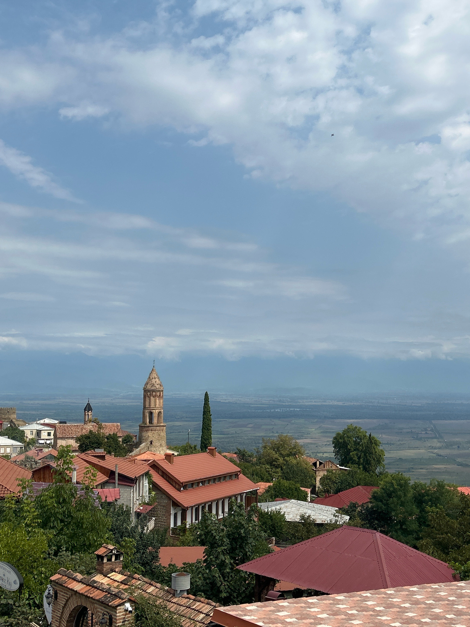 The image depicts a scenic view of the city of Sighnaghi, Georgia, characterized by its distinctive terracotta-tiled roofs and ancient architecture. In the mid-ground, a prominent bell tower, constructed from weathered stone with arched openings, stands tall against a backdrop of rolling hills and a vast, hazy landscape. Several buildings with red tile roofs are visible, interspersed with lush green trees. The foreground is dominated by more tiled rooftops and architectural elements, suggesting a densely populated area with a consistent building style. The sky above is a mix of blue and white clouds, with a small, distant aircraft visible. The overall impression is of a historic town nestled in a picturesque, expansive environment.