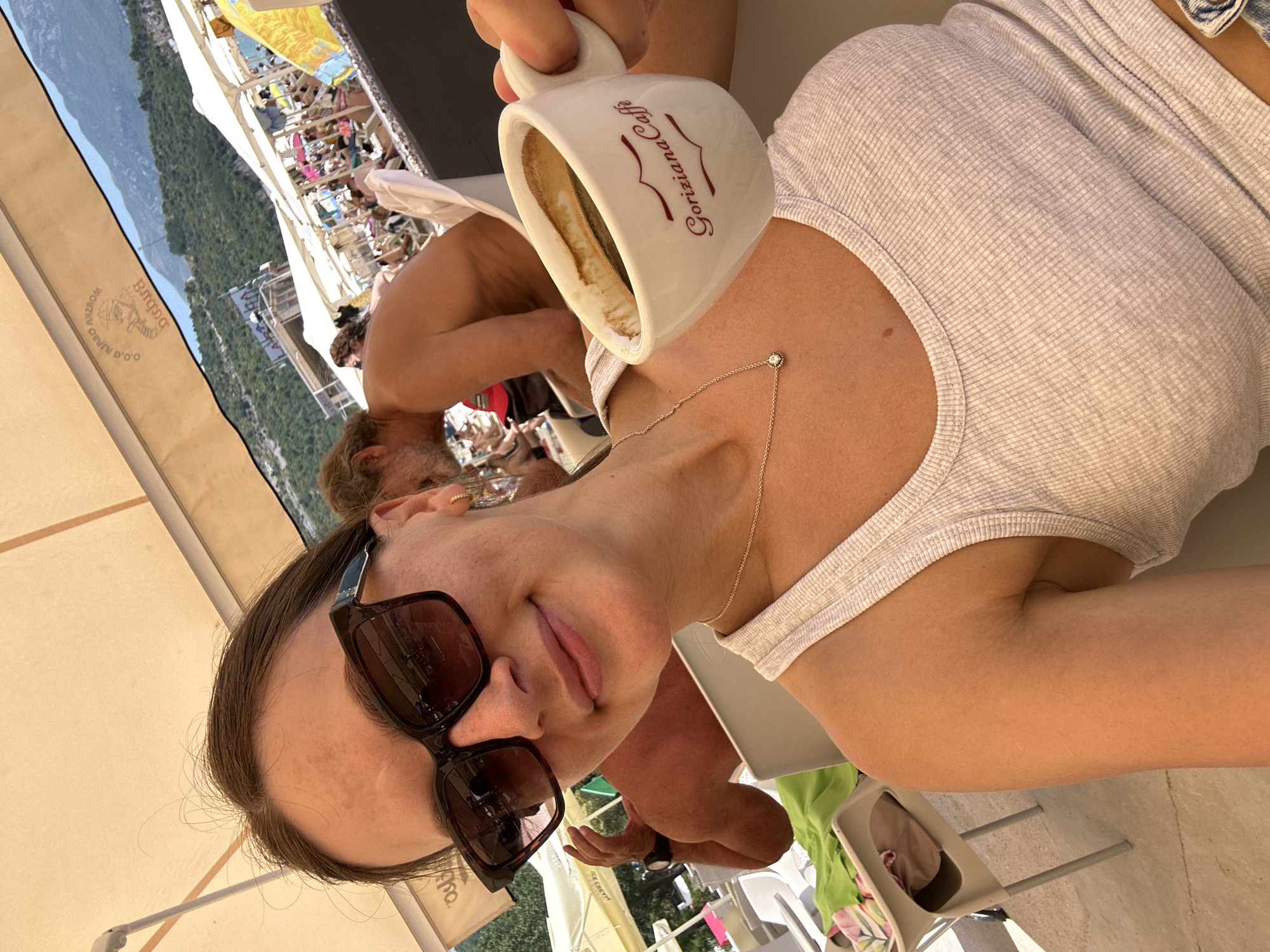 A woman with brown hair, wearing large dark sunglasses, a white tank top, and a delicate silver necklace, smiles at the camera while holding a white coffee cup with "Goriziana Caffe" written in red. She is seated outdoors, possibly at a beach cafe in Budva, Montenegro, indicated by the mountainous landscape and crowded beach scene in the background. Several white umbrellas and beach chairs are visible, along with numerous people enjoying the sunny day. To her left, a man's bare back is visible. The overall atmosphere suggests a relaxed vacation setting.