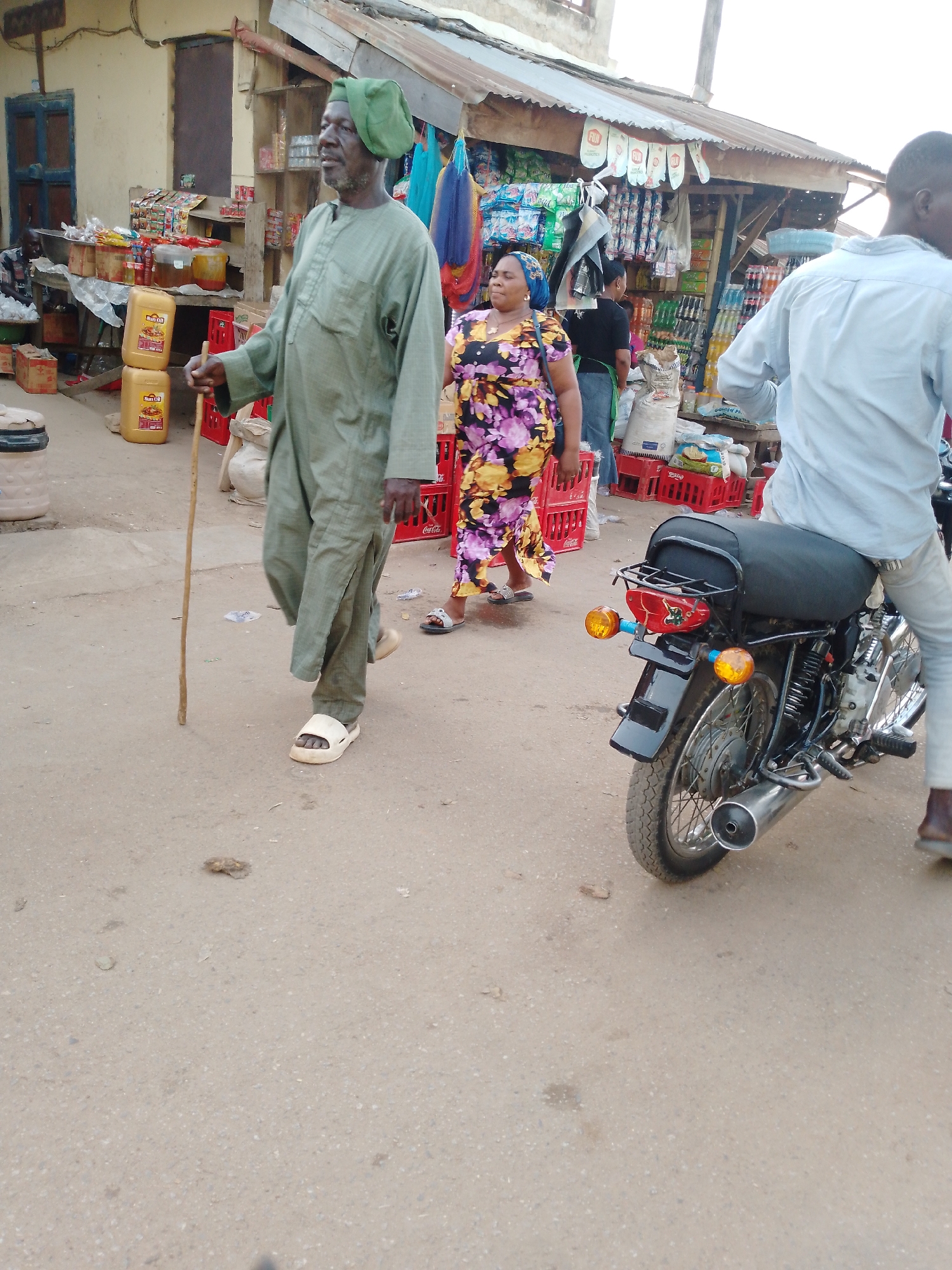 This image depicts a bustling street market scene in Akwanga, Nigeria, during the daytime. In the foreground, an elderly man walks from left to right, dressed in a traditional long green tunic and matching cap, with light-colored sandals. He holds a wooden stick for support and has a visible beard as he looks towards his right. Behind him, a woman in a vibrant floral dress and a blue headscarf also walks in the same direction, wearing light-colored sandals. To the right, a man is seated on a black motorcycle, his back and right leg visible. He wears a light blue long-sleeved shirt and light-colored trousers. The motorcycle features a black seat, chrome exhaust pipe, orange turn signals, and a red taillight.
The background is filled with market stalls under a corrugated metal roof, displaying various goods on wooden shelves. These include packaged food items, bottled beverages, and large white sacks possibly containing grains. Several red plastic crates, some branded "Coca-Cola," are stacked around the stalls. Yellow jerry cans, clearly labeled "Sun Oil," are prominently visible near the elderly man. Small white banners with the word "FUN" are strung along the market roof. A building with a blue door is partially visible behind the stalls. The ground is unpaved, appearing to be a worn dirt or asphalt surface. The scene portrays typical daily commerce and movement in an active local market.