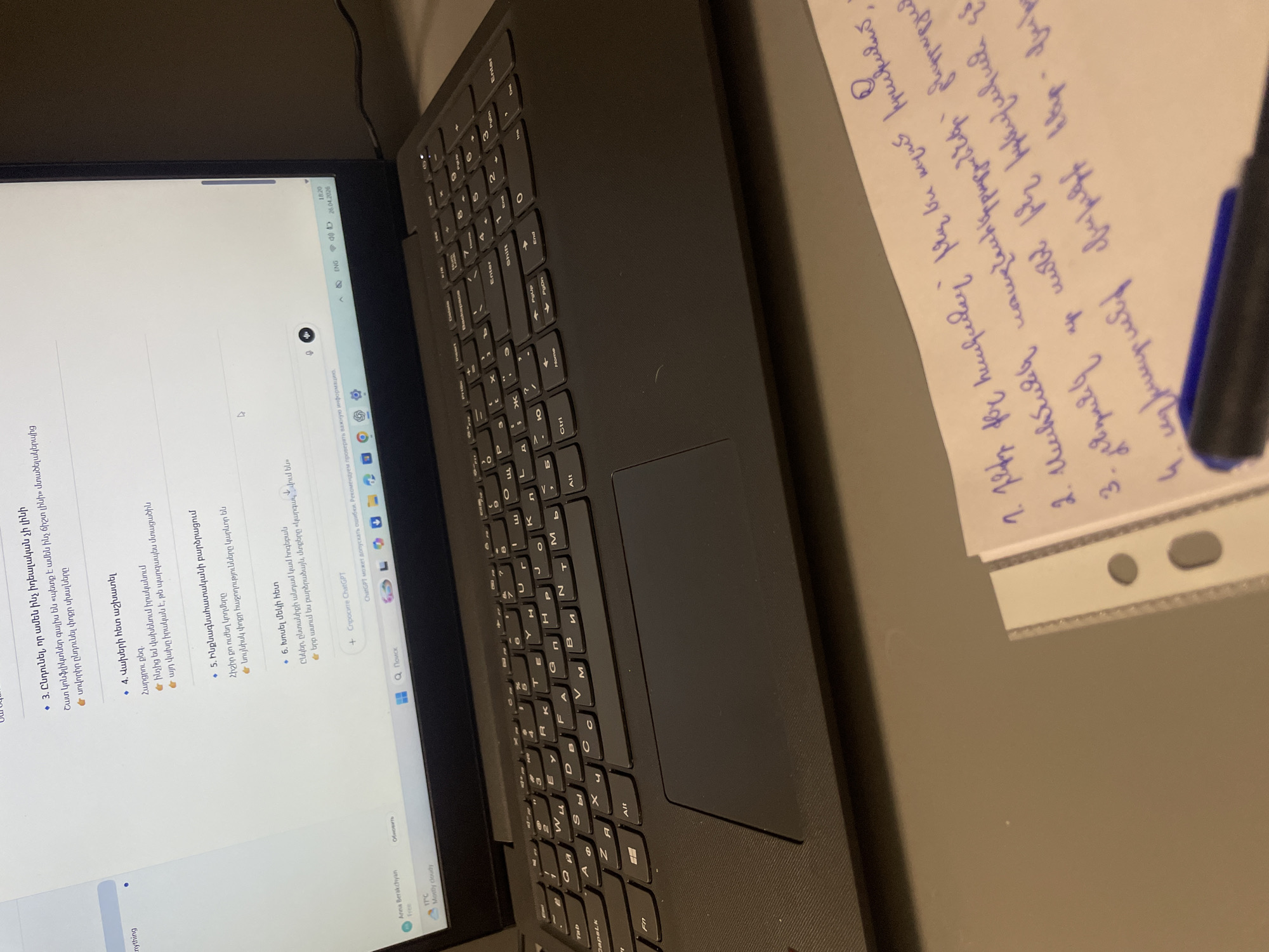 Here is a matter-of-fact description of the scene depicted in the image:
The image shows a laptop and a piece of paper with handwriting on a gray surface. The laptop screen displays text in Armenian, which appears to be related to psychological advice or self-help topics. The laptop keyboard is black, and several keys are visible. To the right of the laptop, there's a white paper with handwritten notes in blue ink, and a blue pen placed on the top of the paper. The angle and lighting suggest the photo was taken indoors. The location is unknown.