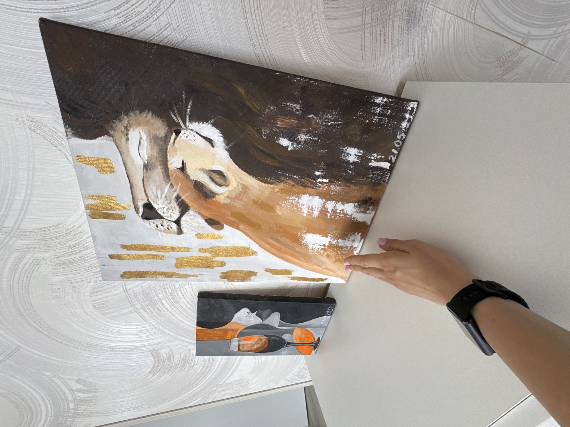 A top-down view showcases two paintings mounted on a wall with textured wallpaper. The larger painting depicts two lions in brown, white, and gold hues. Below it is a smaller, rectangular painting featuring a wine glass motif in shades of gray, black, and orange. A Caucasian person's hand, sporting a black smartwatch, extends towards the upper painting. The paintings are positioned near a white, possibly laminated, structure, which could be a cabinet or wall panel. The location is inferred as Sochi, Russia, based on a signature and date “21.05.2023" on the upper painting.