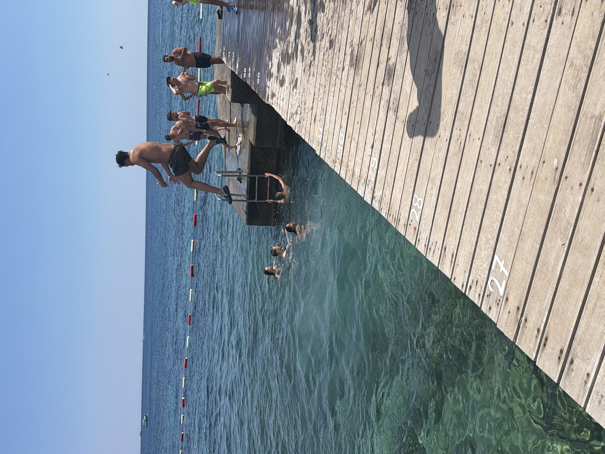 The image depicts a group of young men on a wooden pier by the sea on a sunny day. The scene is set in Budva, Montenegro, as indicated by the location context. The pier, constructed of weathered wooden planks, stretches diagonally across the frame, with numbers painted on some of the planks, suggesting it might be a diving platform or part of a promenade with marked sections.
Several individuals are actively engaged in recreational activities. One person is captured mid-air, in the process of jumping from the pier into the clear, turquoise water. Others are already in the water, swimming or preparing to jump. Some are standing on the pier, watching or waiting their turn, dressed in swimwear. The water is calm and transparent, allowing a view of the seabed in some areas.
The sky is a clear, bright blue, indicative of fair weather, likely midday or early afternoon given the strong sunlight creating shadows on the pier. The overall atmosphere appears lively and energetic, with a sense of fun and enjoyment. The presence of a red and white buoys line in the water further suggests a designated swimming or recreational area. The visible numbers on the pier, like '27' and '28', are the only visible text in the image.
