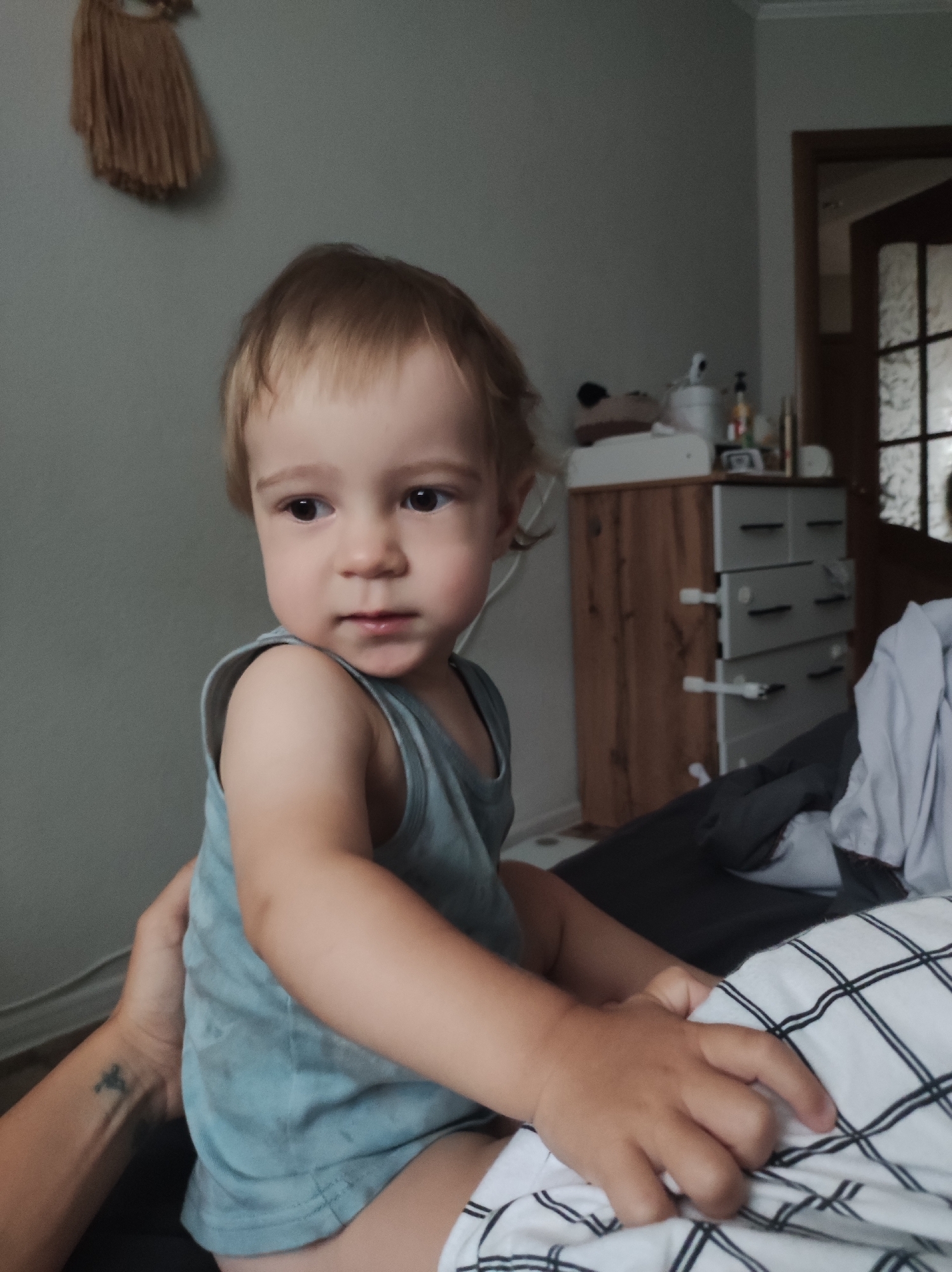A toddler sits on a bed with black and white plaid sheets. The child wears a light blue tank top. An adult arm with a small cross tattoo holds the child. Behind the bed is a wooden dresser with gray drawers, and a door with glass panes. A decorative woven wall hanging is visible in the upper left corner. The photo appears to be taken indoors. There is no indication that this is in Chaikovsky, Russia.