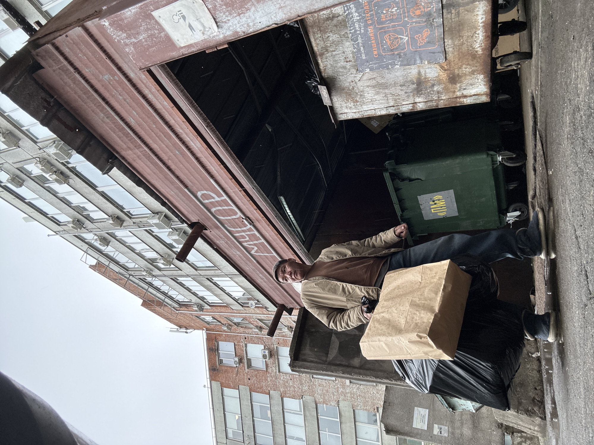 A man stands in an alley next to a dumpster in Khimki, Russia. He wears jeans, a brown shirt, a beige jacket, and dark shoes. He holds a brown paper bag in one hand and what appears to be eyeglasses in the other. The alleyway is surrounded by buildings with windows and air conditioning units. There are black trash bags on the ground. A green trash bin is visible inside the dumpster.