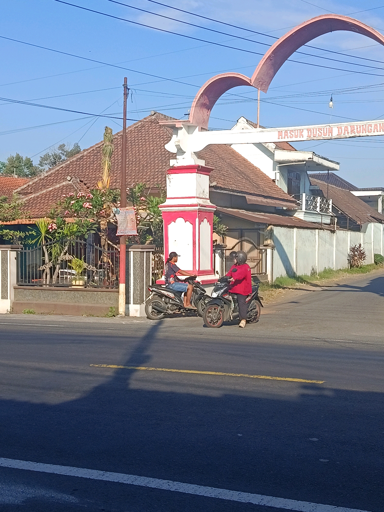 The image shows a street scene in Tempeh Lor, Indonesia, under a clear blue sky. In the foreground, a paved road with yellow and white lane markings is visible, with a long shadow cast across it. Two people are on motorcycles. One person, wearing a blue t-shirt and shorts and a black cap, is riding a dark-colored motorcycle. Next to them, a person in a red top and dark pants, wearing a helmet, is on a similar motorcycle, appearing to be waiting or stopped. Behind the motorcycles, a gateway arch with the text "MASUK DUSUN DARUNGAN" (Entrance to Darungan Hamlet) is prominent. A building with a tiled roof and a decorative pillar is situated near the entrance. A fence with greenery behind it is also visible. Power lines crisscross the upper portion of the image. The overall impression is of a typical daytime scene in a residential area.