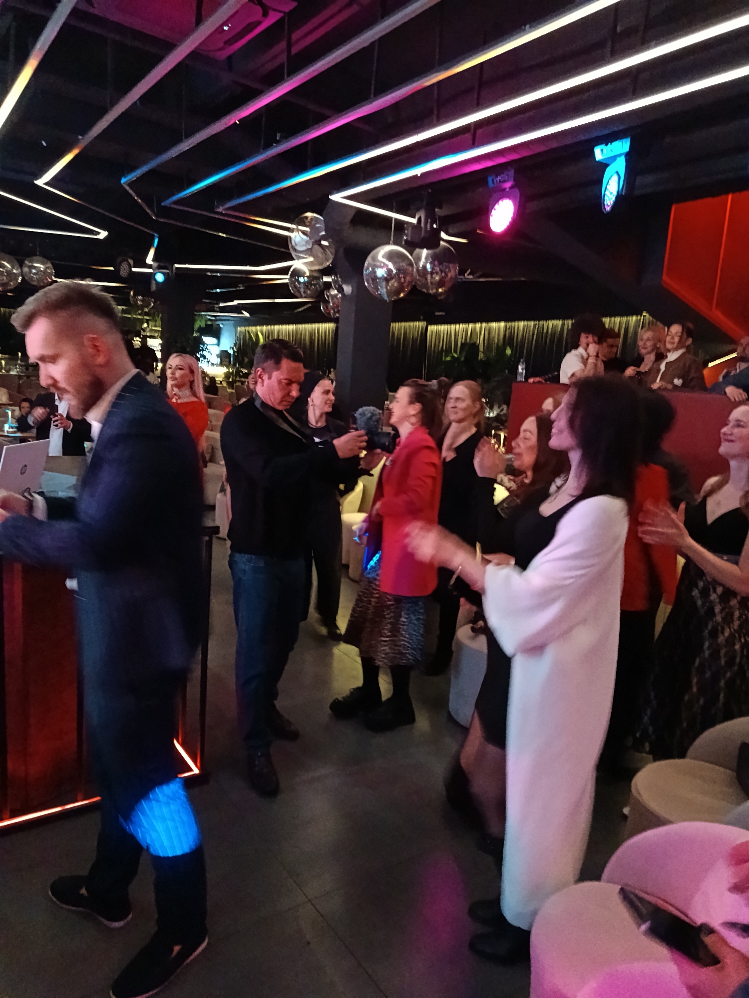 A crowded indoor gathering is captured in an eye-level, full shot. The location has lighting features with mirror balls and neon lighting, providing a modern and festive ambiance. Numerous individuals are present, with some appearing to be clapping or interacting with each other. A man stands prominently in the foreground with an open laptop next to him. Others are holding recording equipment, suggesting a formal event or performance. The people are varied in age and dress, adding to the dynamic and festive setting.