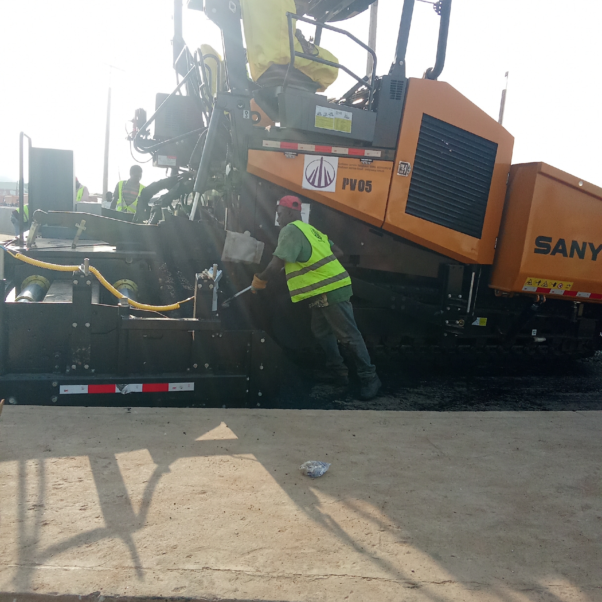 The image shows a road paving project underway in what is reported to be Akwanga, Nigeria. A large, yellow SANY asphalt paver is the central element, attended by a construction worker wearing a green t-shirt, a reflective yellow safety vest, grey pants, and a red cap. He appears to be using a tool to work with the fresh asphalt.
Other construction workers, also wearing reflective vests, are visible in the background. The environment includes a freshly paved section of road, and a piece of discarded trash is visible on the concrete pavement in the foreground.