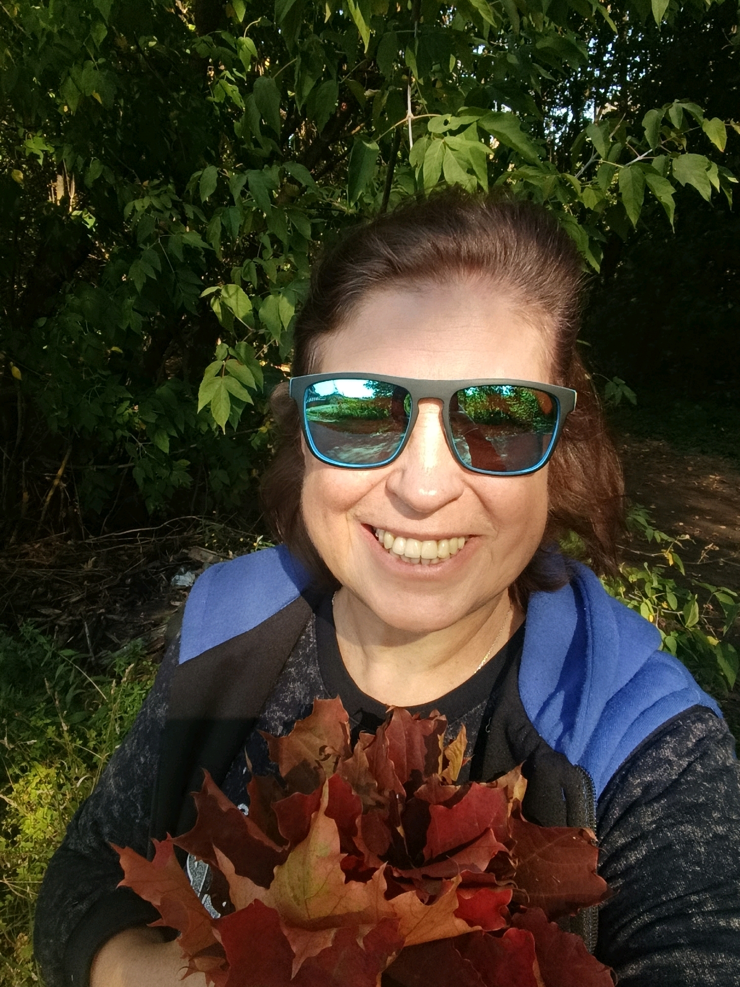 A woman with brown hair and sunglasses smiles at the camera outdoors. She is wearing a blue hooded jacket over a dark shirt and holding a bouquet of red and orange autumn leaves. The background consists of lush green foliage and trees, with sunlight filtering through the leaves. The image appears to be taken during the day, likely in a park or natural area. No specific location in Verra, Russia is identifiable from the image.