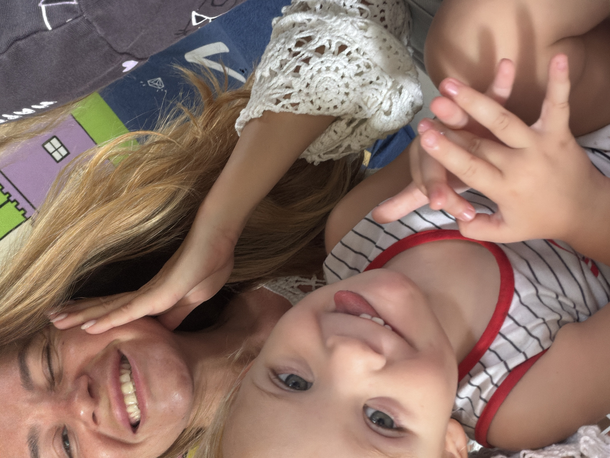 In a close-up, indoor selfie, a woman with long blonde hair and a bright smile winks at the camera while resting her cheek on her hand. Her skin appears tanned and slightly shiny. To the right of her, a young child with blonde hair and blue eyes sticks out their tongue, their hands clasped together in the foreground. The child wears a striped tank top with a red trim. Behind them, a colorful patterned fabric, possibly a bedsheet or blanket, features cartoon-like drawings of houses and geometric shapes in purple, green, and blue. A hint of a dark gray garment is visible on the far right, with white graphic elements. The scene suggests a relaxed, domestic setting, potentially a bedroom or living area. Given the limited visual cues, the precise location within Kuta, Indonesia, cannot be determined.