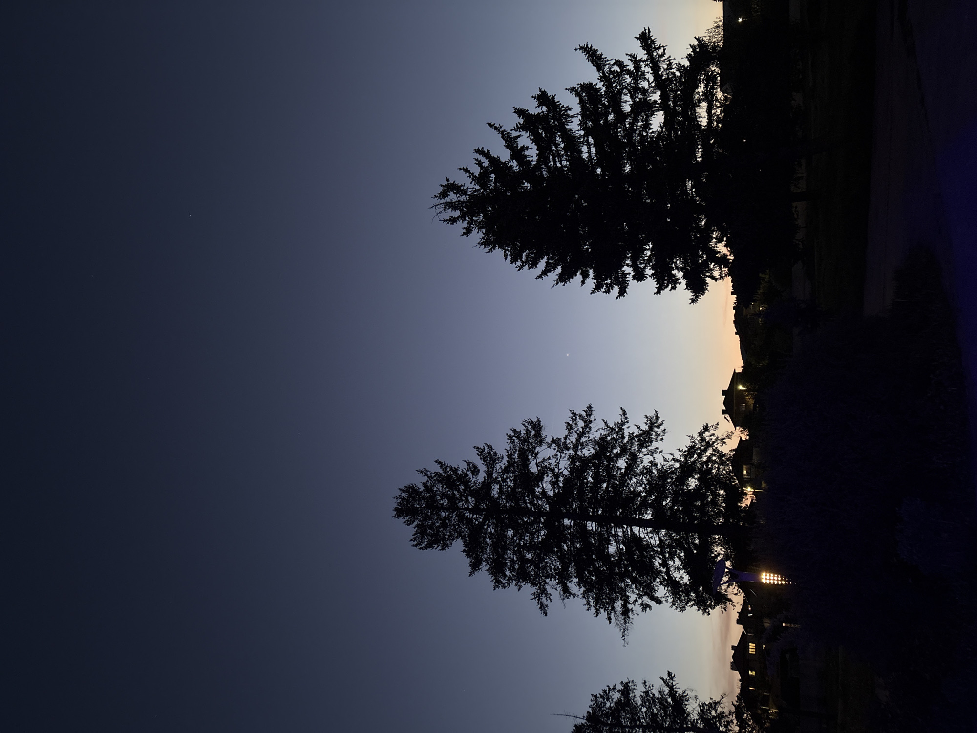 The image depicts an evening landscape with silhouetted trees against a gradient sky transitioning from dark blue to a lighter hue near the horizon. The location appears to be a residential area on the outskirts of a town with buildings and possibly street lights visible in the distance. The overall impression is one of dusk settling over the landscape.