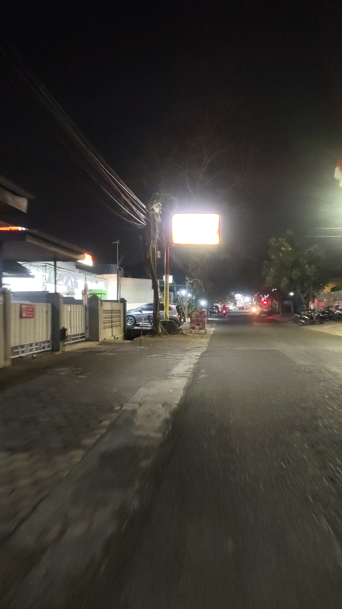 The image depicts a nighttime street scene in the city of Malang, Indonesia. A paved road runs diagonally through the frame, illuminated by streetlights and the headlights of passing vehicles. On the left side of the road, there is a building with a white facade and a fenced-off area. A gray car is parked next to the building. Above the car, a bright, rectangular illuminated sign hangs from a pole, its light casting a glow on the surrounding trees and wires. Further down the road, more lights and vehicles are visible, suggesting a commercial or residential area. Several motorcycles are parked on the right side of the road. The overall atmosphere is dark, with artificial lighting creating pockets of visibility.