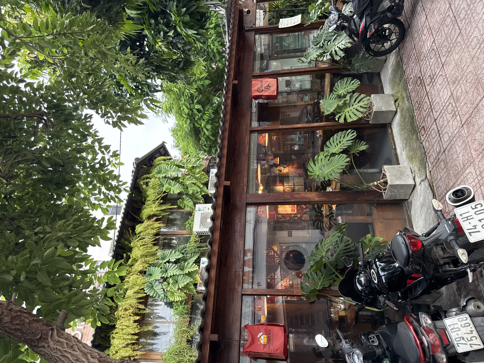 The image depicts the exterior of a restaurant in Nha Trang, Vietnam, on an overcast day. The building features a wooden facade with large glass windows, showcasing the interior where tables and chairs are visible. Lush greenery, including hanging ferns and large-leafed potted plants, adorns the exterior, creating a tropical ambiance.
In the foreground, two motorcycles are parked on a tiled walkway. The motorcycle on the left is red and black, with a license plate reading "79-D1 754.68". The motorcycle on the right is black, with a license plate reading "74-H1 01.05".
A red bag with the text "CHENG AN DIM SUM" is hanging from the building's exterior, near the left side of the image. Another red bag with the text "Cheng An" is hanging on the right side. Air conditioning units are visible mounted above the glass windows. The overall scene suggests a casual dining establishment in a vegetated urban setting.