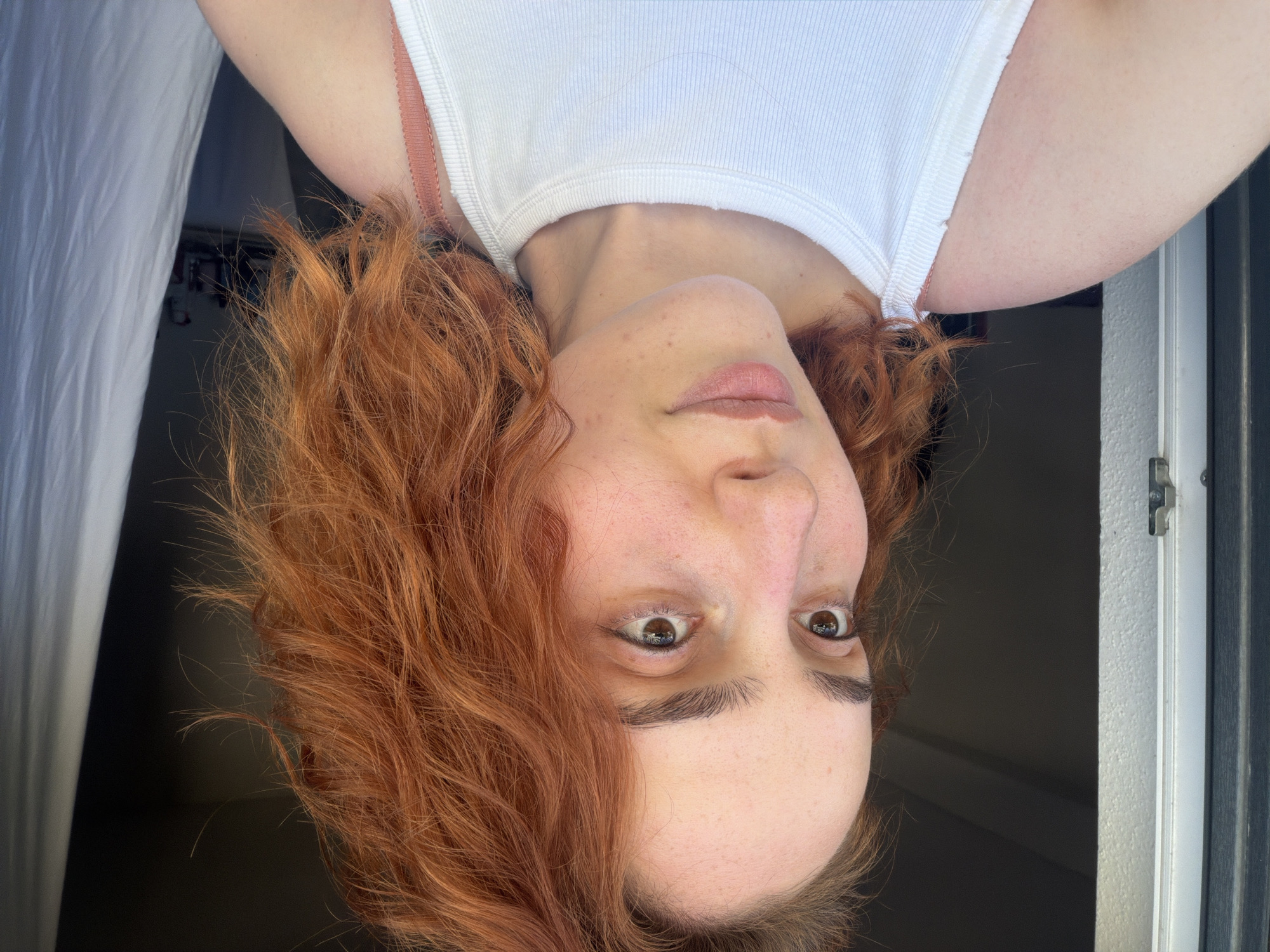 A young Caucasian woman with fair skin, auburn, wavy, shoulder-length hair, and light brown eyes is pictured. She is looking off to her left. She has light dusting of freckles across her nose and cheeks. She is wearing a white ribbed tank top. The background shows an open doorway with a dark interior and a white curtain hanging on the right. The image appears to have been taken indoors, possibly in Strasbourg, France, near an exit leading to an outdoor space.
