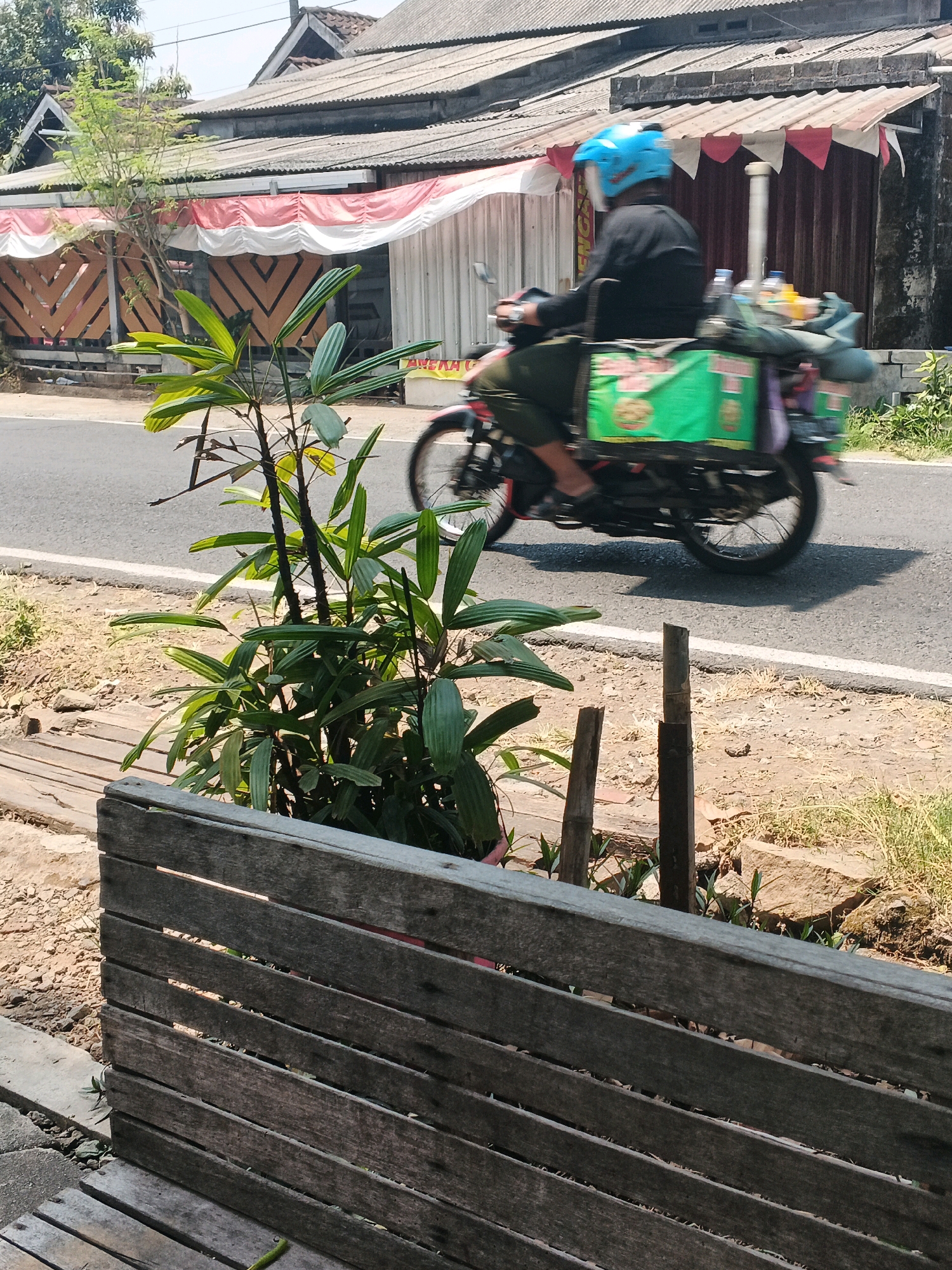 A medium shot captures a street scene in Ungaran, Indonesia. In the foreground, a weathered wooden bench or barrier is visible, constructed from horizontal planks. Beyond this, a paved road runs horizontally across the frame. On the road, a person wearing a black long-sleeved shirt and green pants rides a motorcycle. The rider is wearing a blue helmet and has a green box attached to the back of the motorcycle, filled with various items, including bottles and what appear to be food containers. The motorcycle is in motion, creating a slight blur. To the left of the road, a leafy green plant grows. Behind the road, a building with a corrugated roof is visible, decorated with a red and white striped banner and triangular red and white flags. The building's facade features wooden paneling with a zig-zag pattern. The overall scene is bathed in bright sunlight, suggesting a daytime setting.