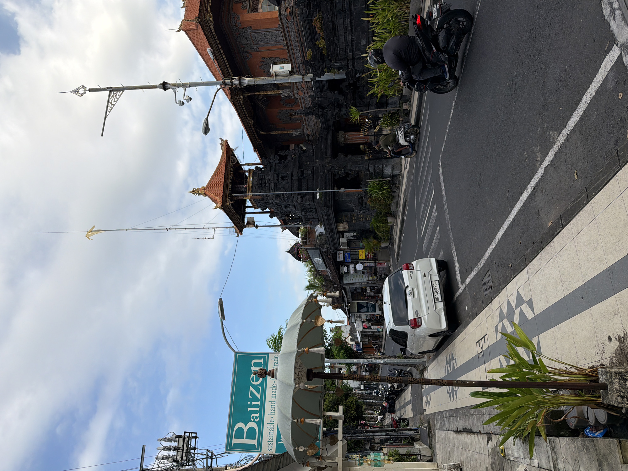 The image shows a street scene in Kuta, Indonesia. A dark-colored traditional Balinese building with a multi-tiered roof dominates the right side of the frame. Parked in front of the building is a black motorcycle. On the left, there is a sidewalk with a "Balizen" sign and an ornamental umbrella. A white car is parked along the sidewalk. Further down the street, other buildings and parked motorcycles are visible. The sky is partly cloudy.