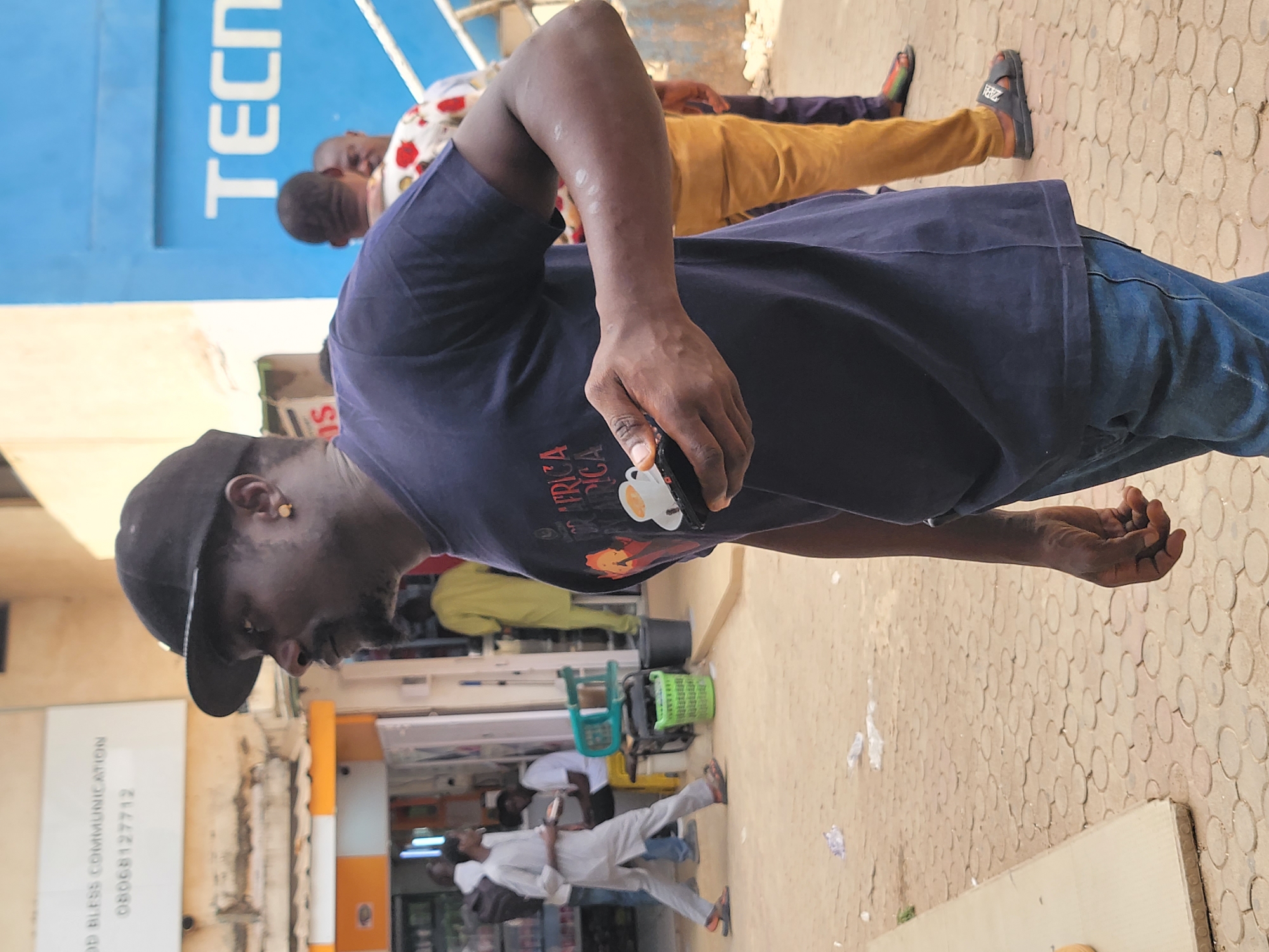 An outdoor daytime scene, likely a street or market area in Jos, Nigeria, features several people walking. In the immediate foreground, a man with dark skin is captured from the waist up. He wears a dark blue t-shirt with a graphic showing two white coffee cups and part of a word, "FRICA". He has a dark baseball cap worn backward and a small gold hoop earring in his left ear. His left arm is bent, and he holds a black smartphone in his left hand. Notable white spots or marks are visible on his left forearm. He is also wearing dark blue jeans.
Behind him, to the left, another man with dark skin is visible, wearing a white shirt with a red floral pattern and tan/yellow trousers, with dark slippers. Further back on the right, a man in a light-colored (possibly white or light grey) traditional tunic and trousers walks while holding a small device. Other blurred figures suggest a busy environment.
The ground is paved with light-colored interlocking bricks, many featuring a circular pattern. In the background, buildings with shopfronts are visible. A prominent bright blue sign with large white letters spells "TECNO". To the far left, a white rectangular sign has black text that reads "GOD BLESS COMMUNICATION" followed by a phone number "08068127712". Green and black plastic baskets or crates are also seen on the ground near the background figures. The bright, even lighting indicates it is daytime with clear weather.