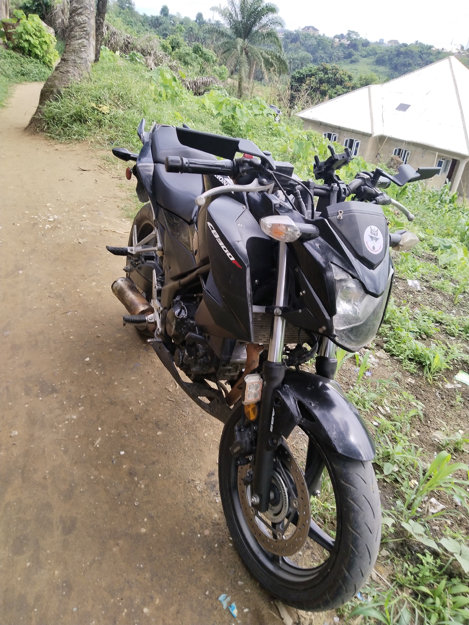 The image depicts a black Honda CB300F motorcycle parked on a dirt path in Uyo, Nigeria. The motorcycle is the primary subject, positioned at an angle with the front wheel prominently displayed. The background features lush green vegetation and a house with a distinctive white, pyramid-shaped roof. The dirt path indicates a rural or semi-urban setting, likely on the outskirts of the city. There are no people visible in the scene. The focus is on the motorcycle and its immediate surroundings, providing a snapshot of a vehicle in a specific geographical location.