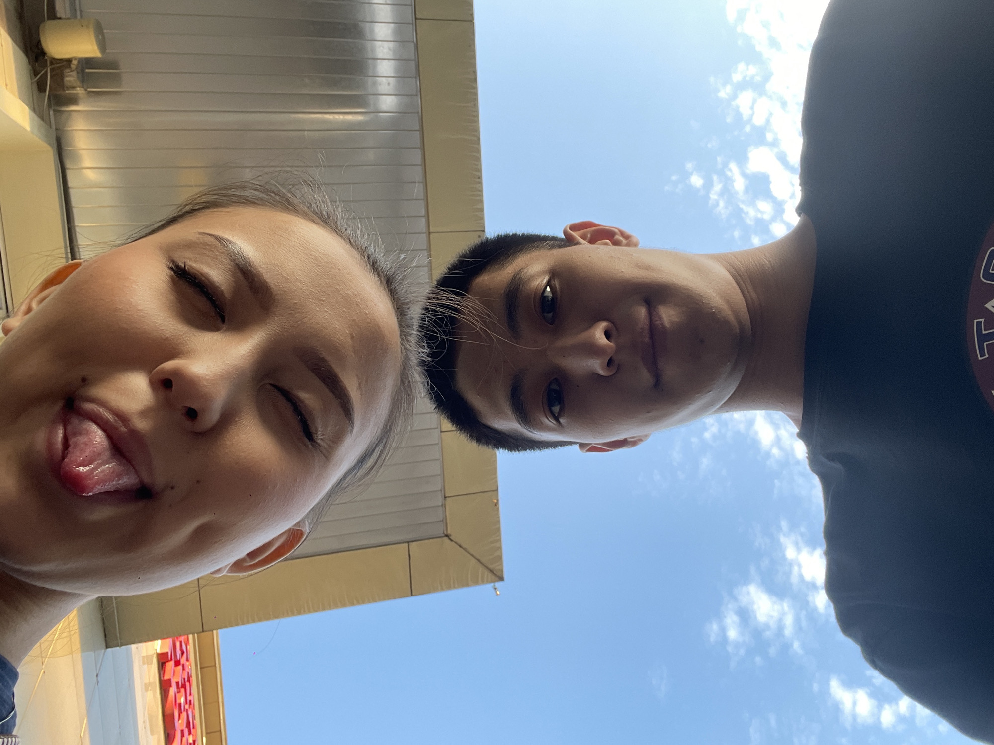 The image shows a close-up, low-angle selfie of two individuals, a woman and a man, taken outdoors during the day. The woman's face is positioned at the top of the frame, upside down, with her eyes closed and her tongue sticking out playfully. She has makeup on her eyes and appears to be wearing a dark top. The man's face is below hers, right-side up, looking directly at the camera with a slight smile. He has short dark hair and is wearing a dark t-shirt. Behind them, the sky is visible with scattered white clouds. Above the man's head, a portion of a building's overhang is visible, appearing to be a yellow awning with metal paneling. The scene suggests a casual outing, possibly in a public space within the city of Almaty, Kazakhstan, given the architectural style which is consistent with some commercial buildings in the area.