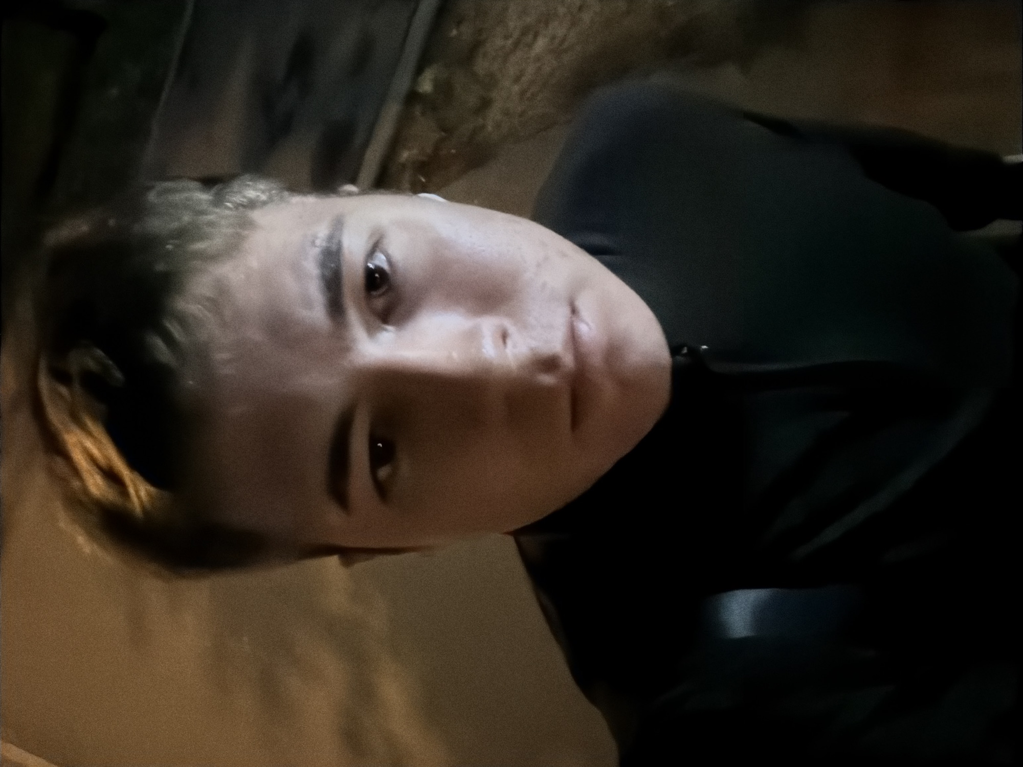 A high-angle, close-up, selfie-style shot shows a young Caucasian male facing the camera. He has short, light brown hair, light-colored skin, and appears to be in his late teens or early twenties. His eyes are dark brown. He is wearing a dark-colored, long-sleeved shirt with a zipper at the neck. A white wireless earbud is visible in his right ear.
The background is dimly lit and appears to be outdoors at night. To the left of the subject, there is a blurred area of a light brown wall or pavement. To the right of the subject, the ground is covered with what looks like gravel or dirt, with some dry leaves visible. A dark, rectangular object, possibly a curb or a raised platform, runs along the right edge of the frame. The overall lighting suggests an urban environment at night, with artificial lights creating subtle highlights on the subject's face and hair. The image quality is grainy, indicating it was likely taken in low light conditions.
The image's metadata does not contain information to confirm its location in the city of Khimki, Russia.