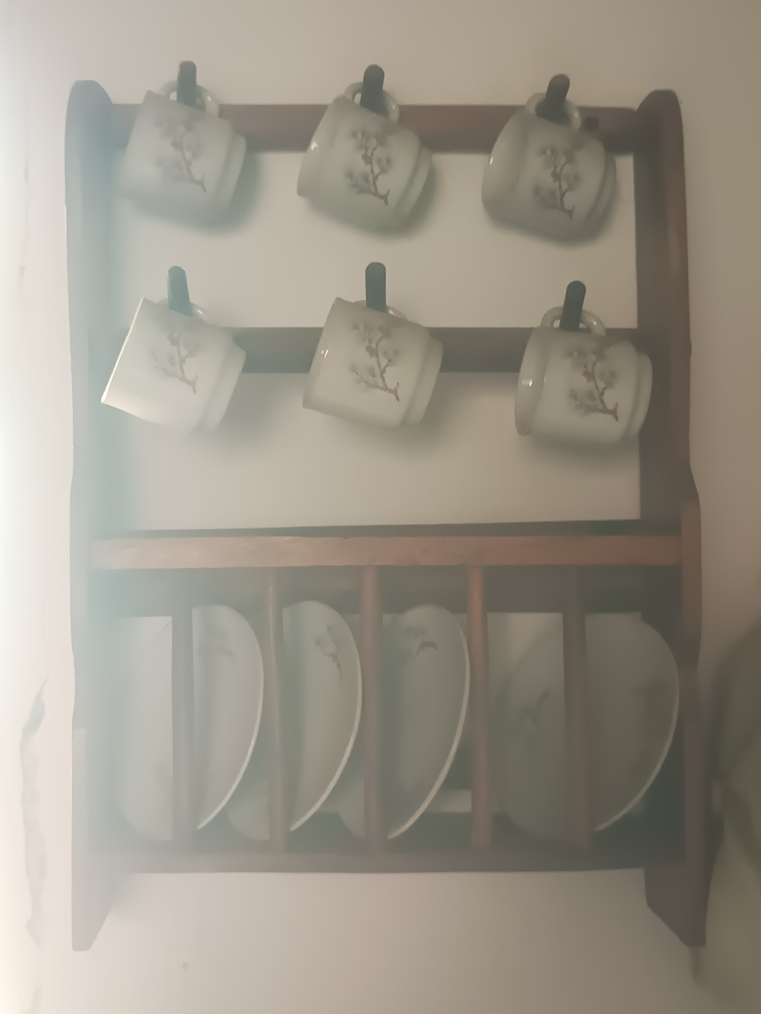 The image shows a wooden dish rack mounted on a white wall. The dish rack has two shelves. The top shelf holds six ceramic mugs, three on the left and three on the right. Each mug is white with a delicate floral pattern. The mugs are hung upside down from hooks. The bottom shelf holds six ceramic plates, also arranged three on the left and three on the right. The plates are white with a similar floral pattern. The scene is well-lit, with a bright light source coming from the left side of the image, casting a soft glow and creating a slightly hazy atmosphere. The dish rack and its contents appear to be in good condition. The overall setting suggests a domestic environment, possibly a kitchen or dining area, in Juan Griego, Venezuela.