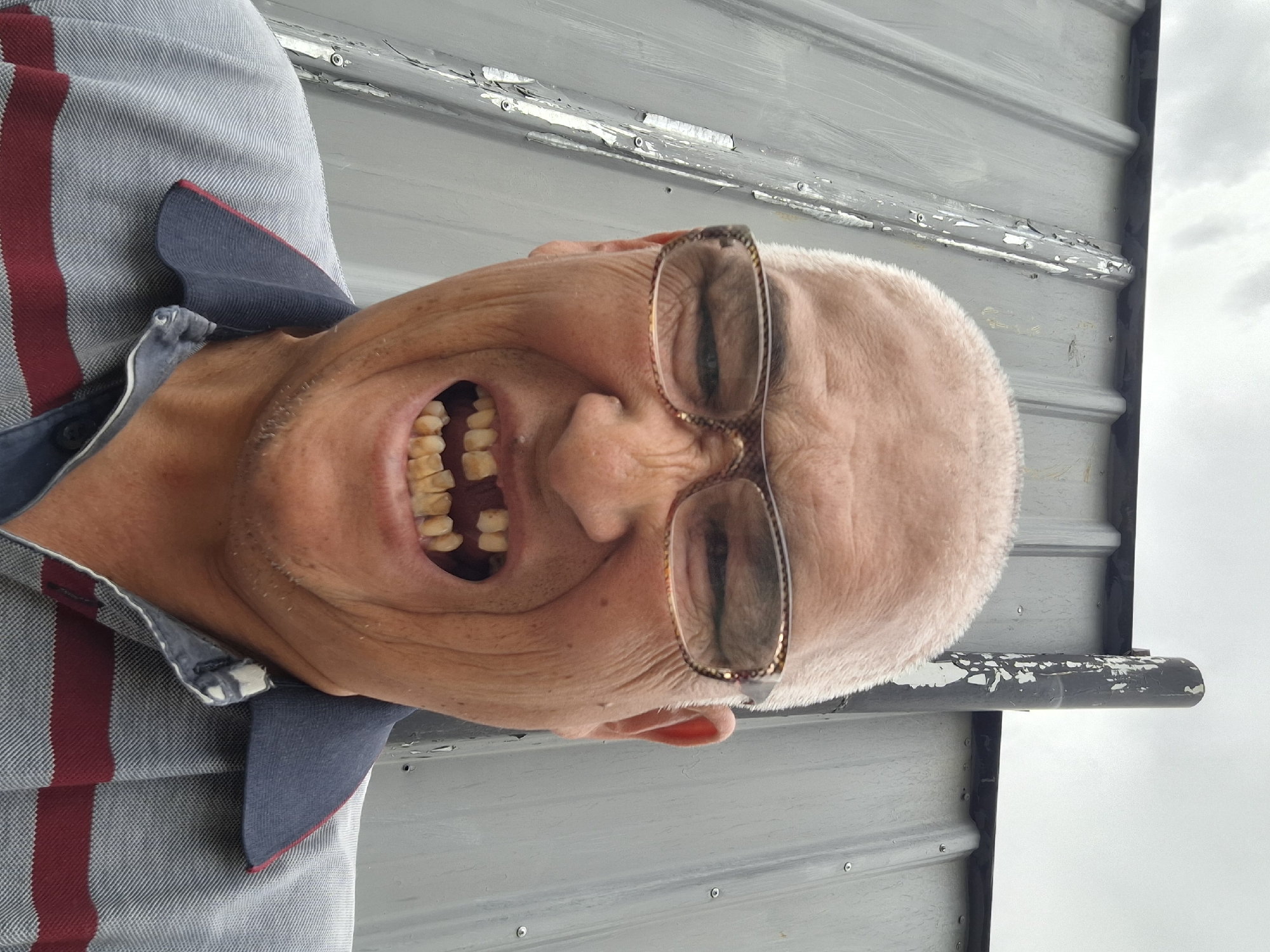 A close-up, low-angle, selfie-style shot shows an older Asian man with a shaved head and glasses. He is wearing a grey polo shirt with red and blue stripes. The man is grinning widely, revealing missing and discolored teeth. He is standing in front of a corrugated metal wall. A dark, cylindrical pipe rises vertically to his right. The sky is overcast. There are no discernible landmarks that would allow for precise location identification in Almaty, Kazakhstan.