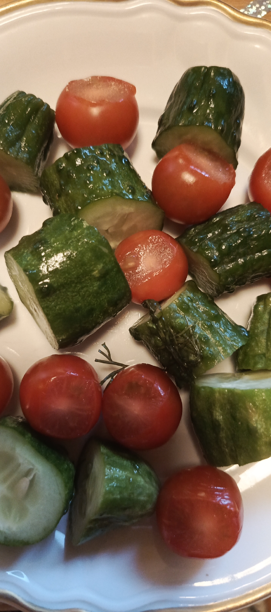 A close-up, overhead shot of a white plate filled with chopped cucumbers and cherry tomatoes. The cucumbers are cut into irregular chunks, revealing their green, bumpy skin and pale interior. The cherry tomatoes are whole or halved, their red skin glistening. Some of the tomatoes have been cut open, exposing their juicy, seed-filled flesh. The food is arranged in a somewhat haphazard manner on the plate, with no discernible pattern. The background is a blurred, golden rim of the plate, and the surface beneath it, suggesting a table setting. There is no information about the location in the city of Мерчалово Снт, Russia, present in the image.