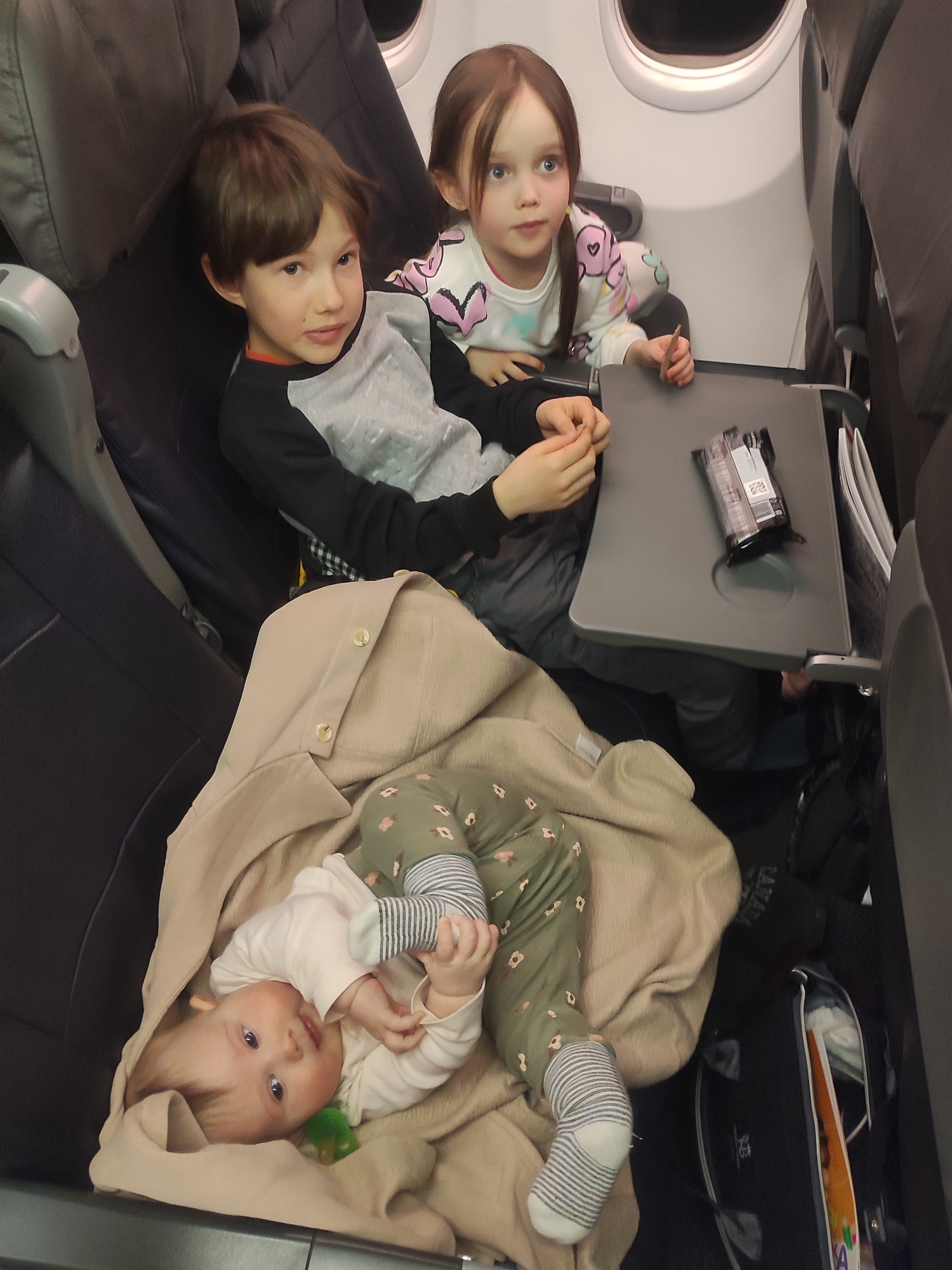 This image depicts the interior of an airplane cabin. The scene does not contain visual identifiers specific to the city of Sochi, Russia; rather, it portrays a general airplane environment.
Three children are present in the frame. In the center, a young boy with brown hair, wearing a black and gray long-sleeved shirt, sits in an airplane seat, looking directly towards the camera. To his right and slightly behind him, a young girl with long brown hair tied in pigtails, wearing a white long-sleeved shirt with colorful patterns, sits by a window (porthole). She holds a light-colored, elongated object in her right hand and gazes towards the right side of the frame, away from the camera. The tray table in front of her is deployed, holding a dark-colored packaged snack. In the foreground, spanning the lower half of the image, an infant is lying on a light tan blanket. The infant is wearing a white long-sleeved shirt and green patterned pants with striped socks, looking upwards towards the camera with a green pacifier near its mouth. The blanket covers the infant's lower body and is draped over what appears to be another airplane seat. A black bag is visible on the floor to the right of the infant. The airplane seats are dark-colored. The cabin lighting appears dim, consistent with an in-flight setting.