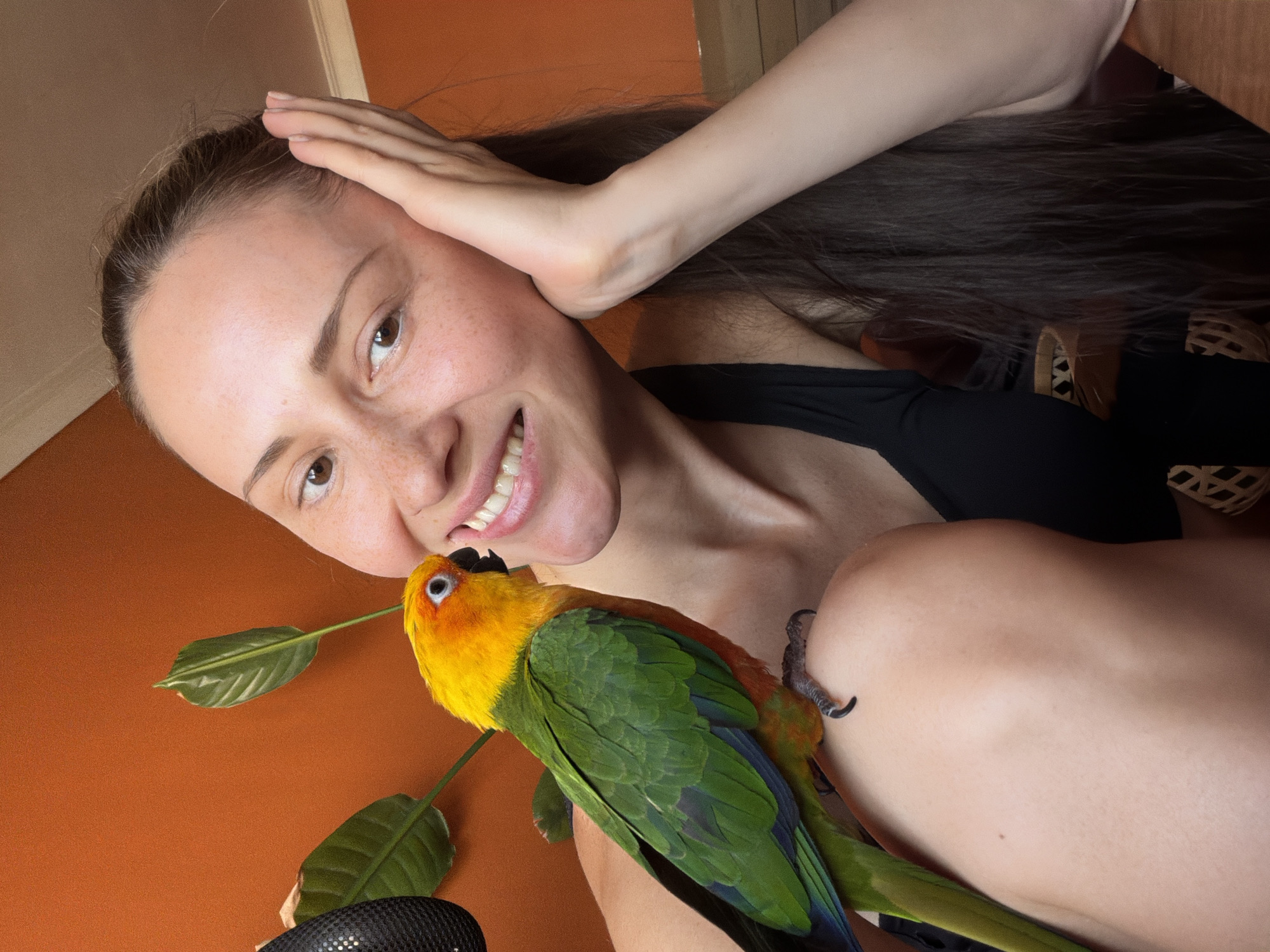 A person, presumably female, is holding a brightly colored parrot. The person's face is visible with a smile, and their hand is on their head. The parrot is perched on the person's shoulder, holding onto a leafy green stem. The background is a solid orange color. There is no indication in this image that the location is Khimki, Russia.