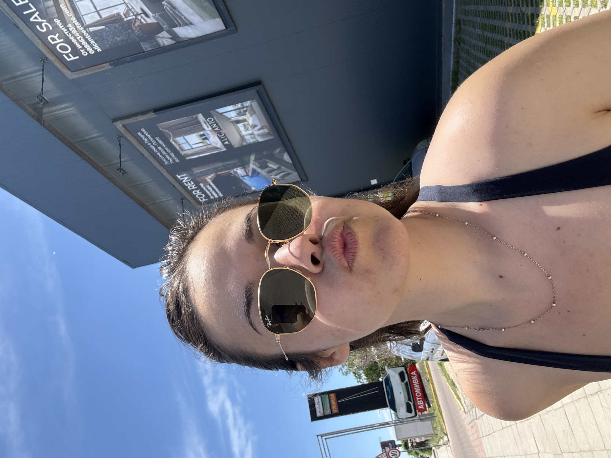 The image is a selfie taken outdoors on a sunny day, featuring a person wearing sunglasses and a dark top. Behind the person is a building with signs, including one that reads "FOR RENT" with the text "ALICANTO" and a description in Bulgarian about a luxury house in Greece. Another sign says "FOR SALE" and includes the website "alicantoestate.com" and a phone number. In the background, there's a street sign written in Cyrillic (АВТОМИВКА), a white vehicle, and other street signs including speed limit signs. Based on the Cyrillic text, the location is likely in Bulgaria. Given the signs for "ALICANTO" and "alicantoestate.com", it is near the "Alicantoestate" real estate office in Sofia. The weather is clear and sunny, indicating it might be during the summer months.