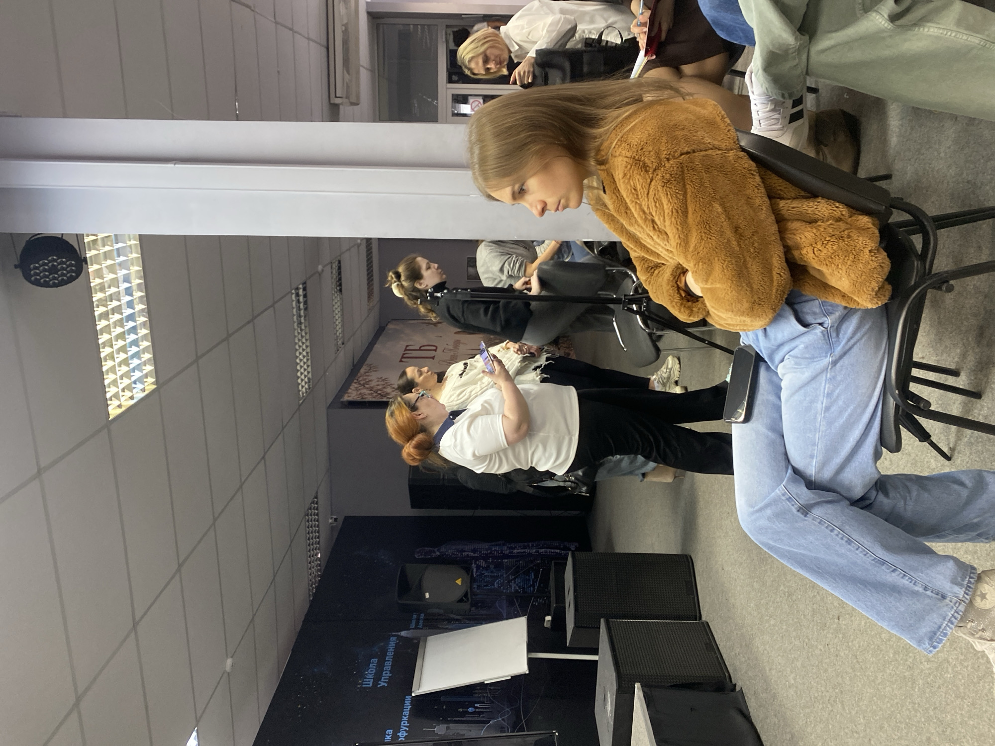 Here is a detailed description of the image:
The image depicts a gathering of people indoors, possibly in an educational or conference setting. There are several individuals seated on chairs, their attention seemingly directed towards a presentation or speaker.
The foreground is dominated by a young woman in a brown, fluffy jacket and blue jeans. She is seated on a black chair and appears to be looking down. Other individuals can be seen in the background, some using their phones.
A white board and several black speaker boxes are in the foreground, suggesting a presentation or lecture is taking place. A large screen in the background has some text and an image displayed.
The room appears to have fluorescent lighting on the ceiling and a gray wall. The environment suggests a modern, functional space meant for presentations, lectures, or meetings. The location details, including the city and specific place, are not readily discernible from the image.