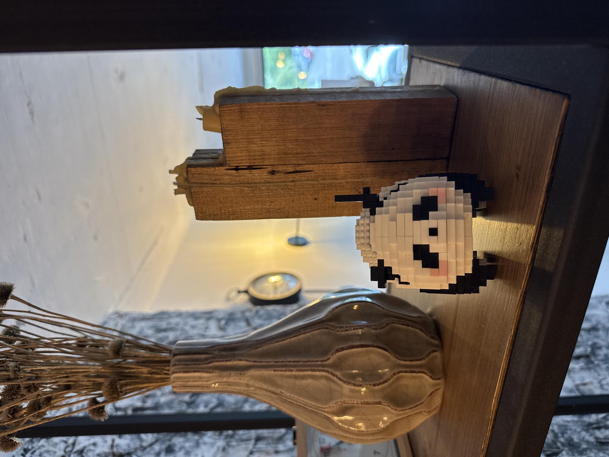 The image depicts a shelf with various decorative items. On the left, a tall, beige, textured vase holds dried, spiky foliage. To the right of the vase, a small, pixelated panda figurine is placed on the shelf. Behind the panda, two tall wooden blocks are positioned, with remnants of melted wax from candles visible on top of them. In the background, a clock with a dark frame and a circular face is mounted on a light-colored wall. The lighting suggests an indoor setting, possibly a home or a shop, with a warm yellow light source illuminating the area. The overall aesthetic is rustic and decorative. This image does not provide any information about the city of Batumi, Georgia.