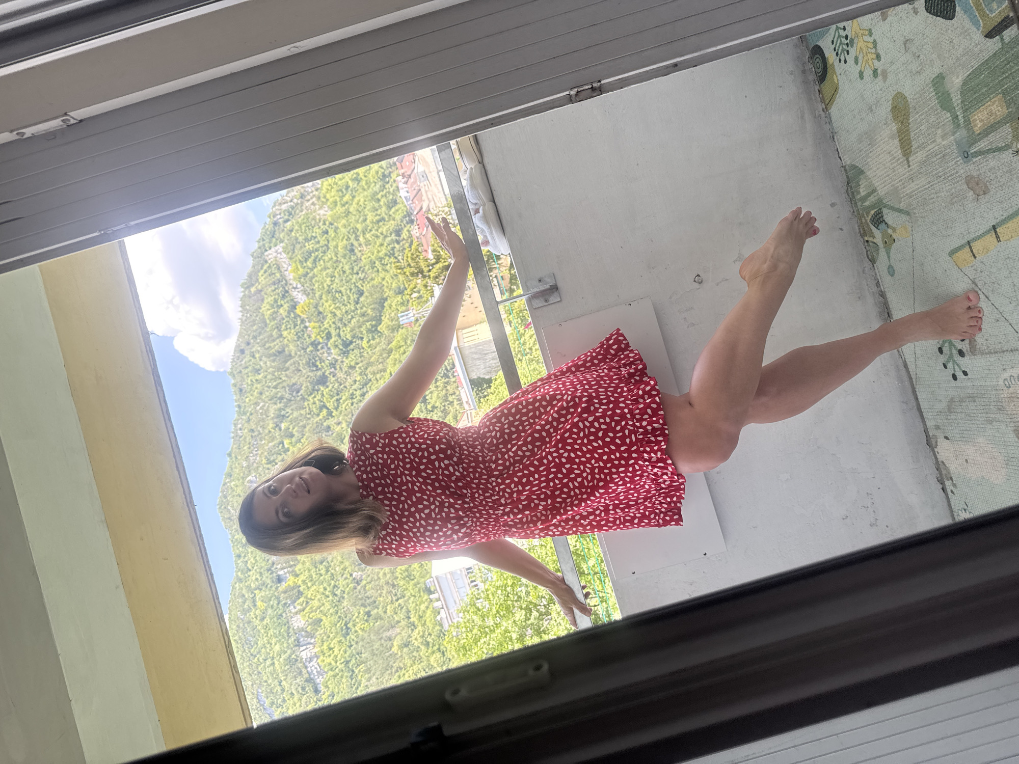 A woman in a red, white-spotted dress is posing on a balcony with a mountainous, tree-covered landscape in the background. The woman is barefoot and has her arms extended. Her left leg is bent, and her right foot is lifted off the ground. She appears to be in Saint-Claude, France, as evidenced by the architecture visible in the distance. The balcony railing is silver, and there are two pairs of white shoes on the railing. The floor of the balcony is covered with a mat with a pattern of green and yellow shapes. The image is framed by a dark doorway on the left and right.