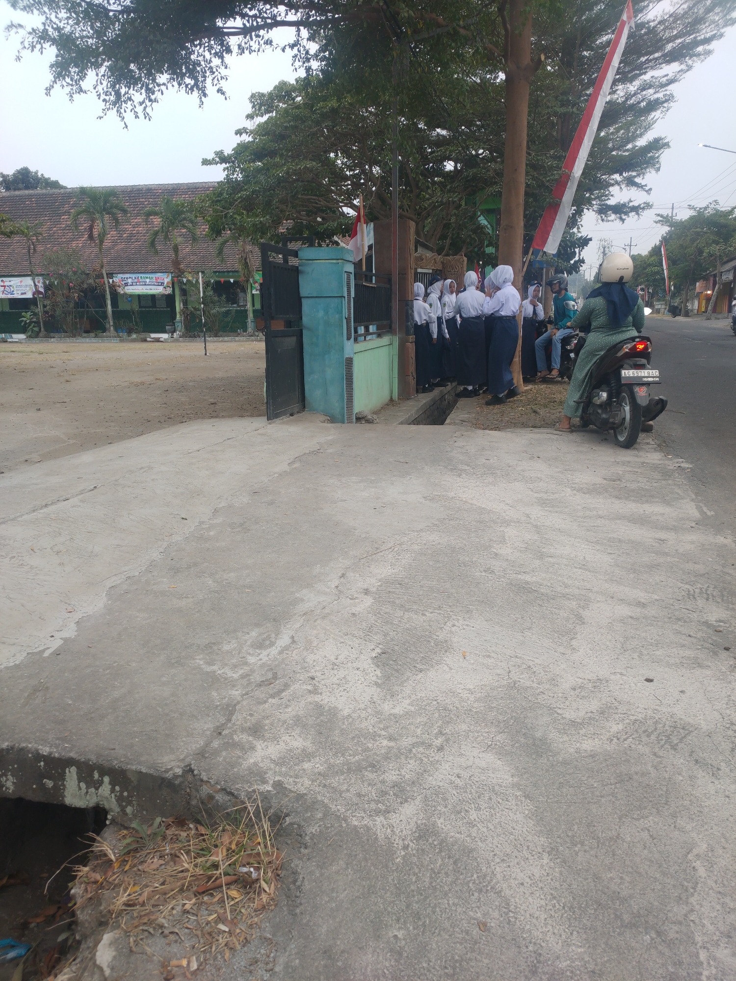 In Banggle, Indonesia, a group of students in white and navy uniforms are gathered near a school entrance. A woman wearing a green patterned dress and a white helmet is seated on a dark-colored scooter. In the background, a building with a red-tiled roof and palm trees is visible. The scene is observed from a slightly elevated concrete walkway, which slopes down to a ditch filled with dry leaves and grass. A tall tree with lush green foliage stands prominently, partially obscuring an Indonesian flag that is attached to a pole. The sky is overcast.