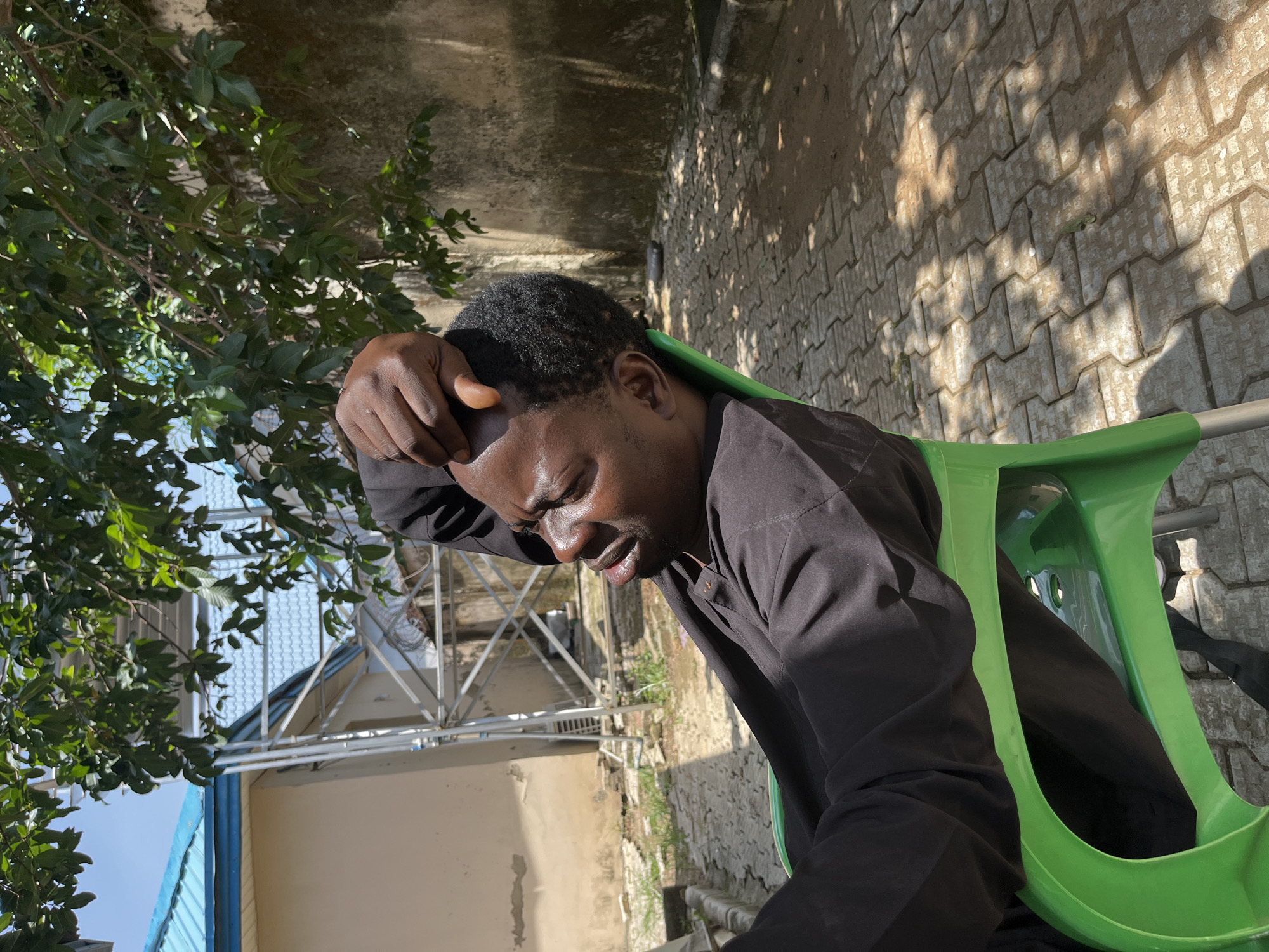A man is sitting in a green plastic chair outside, leaning back with his head tilted up and his eyes closed. He is wearing a black collared shirt. His right hand is resting on his forehead. The ground is paved with interlocking bricks. In the background, there is a wall, some trees with green leaves, and a building with a blue roof. A metal structure, possibly scaffolding or a tower, is also visible. The scene is lit by sunlight, casting shadows on the ground. The location is identified as Galadimawa Village, Nigeria.