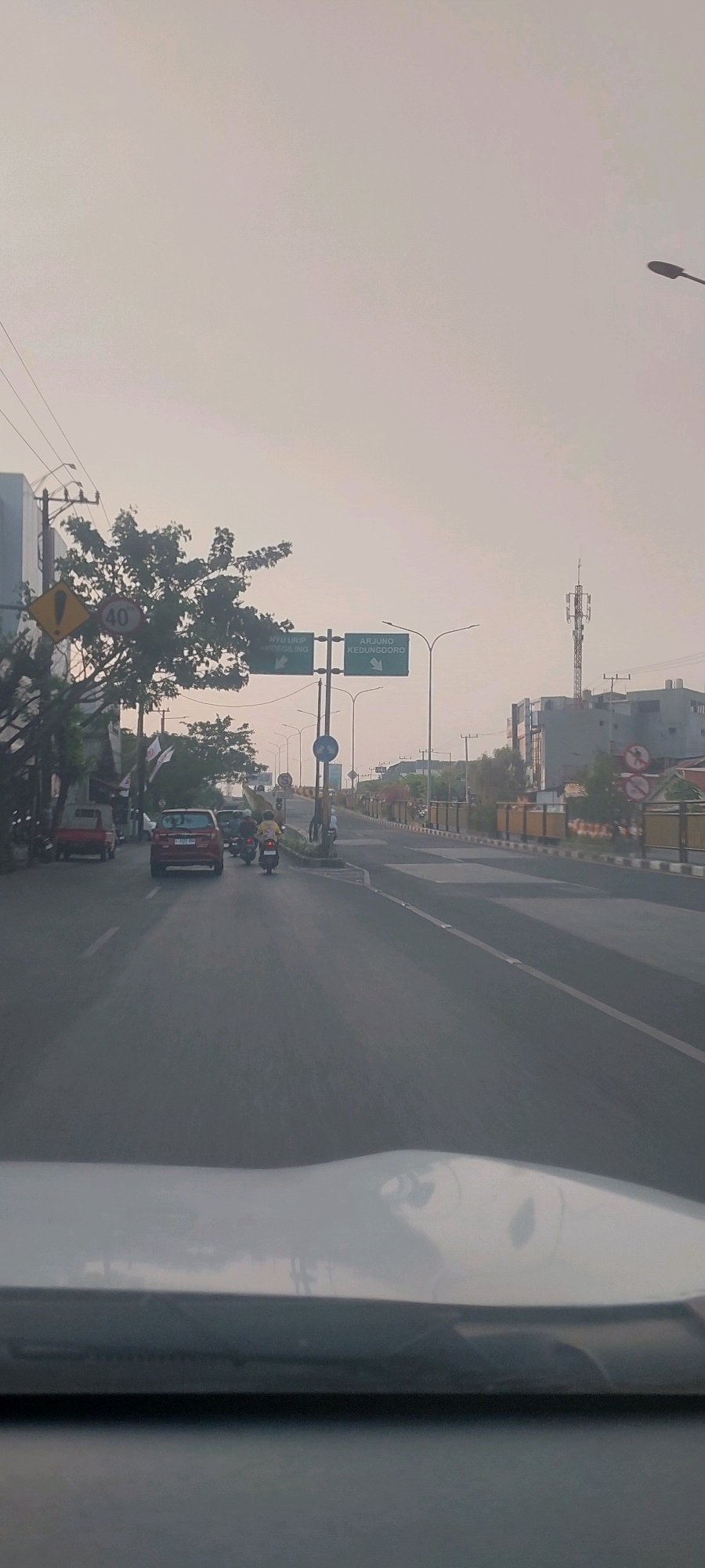 The image depicts a street scene in Surabaya, Indonesia, under an overcast sky. In the foreground, the dashboard of a vehicle is visible. The road is paved and appears to be a multi-lane street. Traffic is present, including several cars and motorcycles. On the left side of the road, there are buildings, trees, and power lines. Road signs are also visible, including a directional sign pointing to "ARJUNG KEDUNGDOORO" and another indicating "NYU URIP / KRONGSONG". A speed limit sign showing "40" is also present. On the right side of the road, there is a sidewalk with a fence and some buildings. A cell tower is visible in the distance. The overall atmosphere is calm, suggesting it might be early morning or late afternoon.
