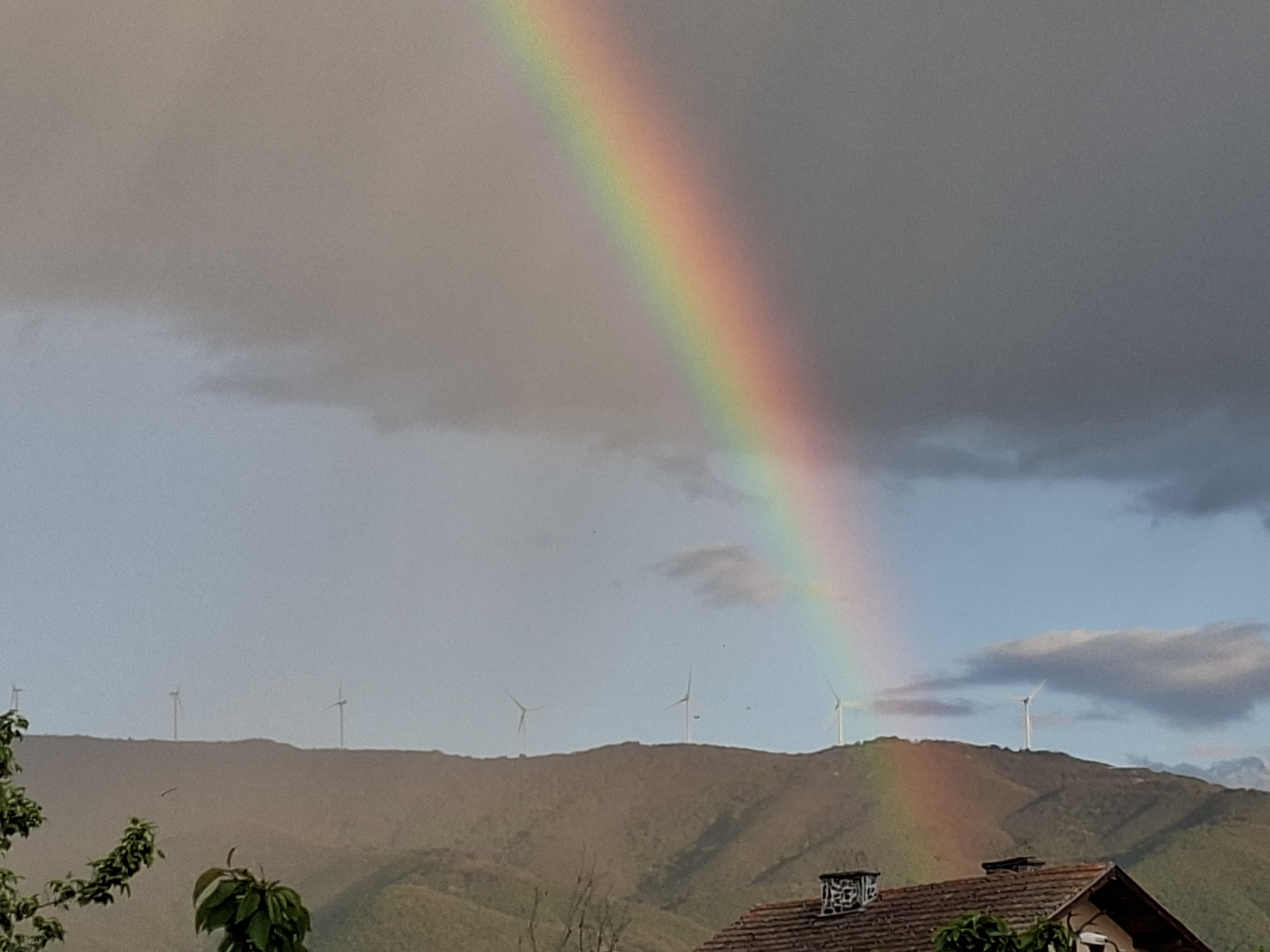 The image shows a rainbow arcing across the sky. In the foreground, there is a tiled roof of a building. Beyond the roof, a mountain range is visible with several wind turbines positioned along the ridgeline. The sky transitions from cloudy on the left to a clearer blue on the right side of the rainbow. The location is Unknown, Unknown.