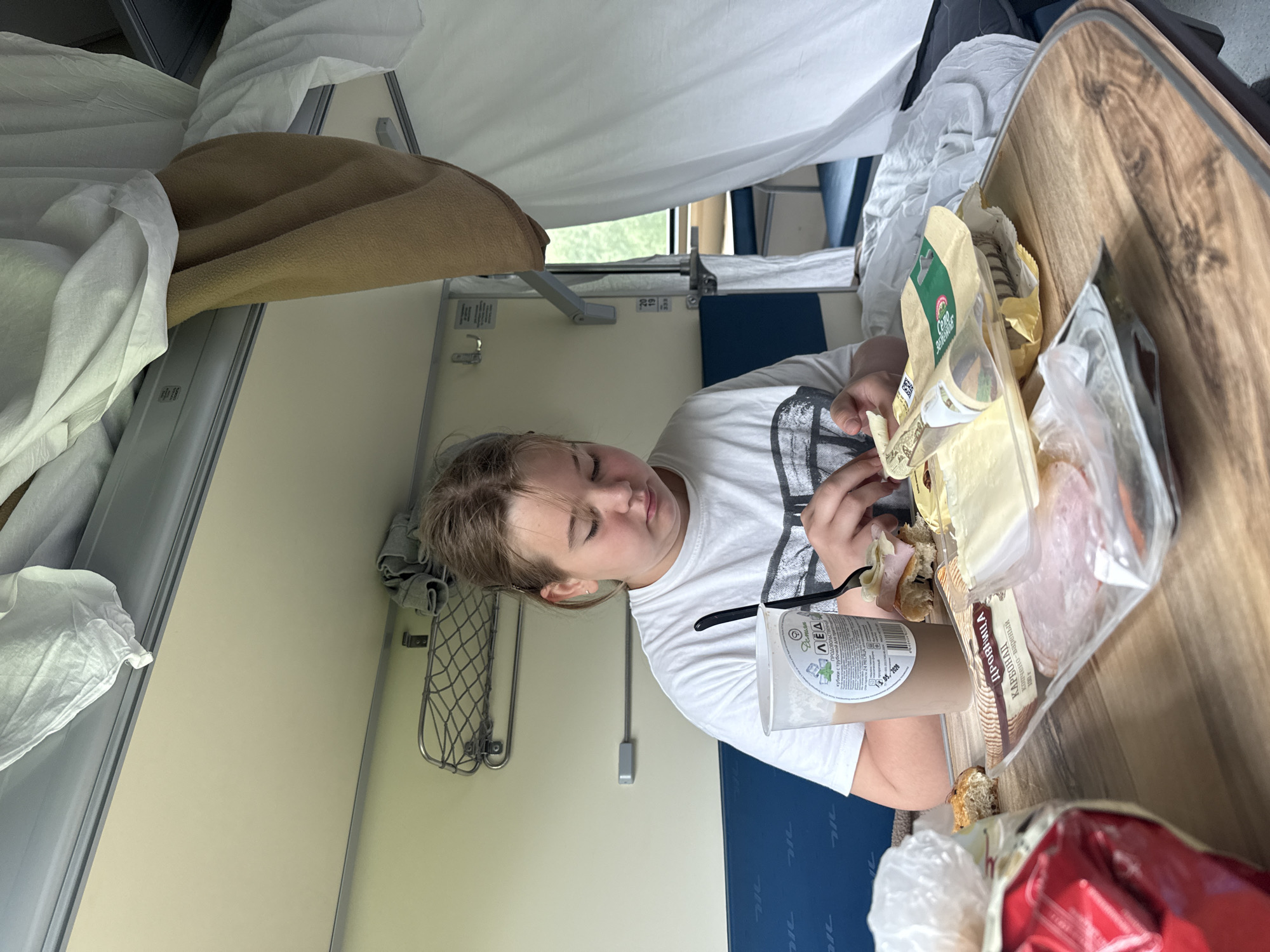 A young person is sitting at a wooden table inside a train compartment. The person is wearing a white t-shirt and is focused on preparing a sandwich with ham and cheese. There are food items on the table, including a drink in a plastic cup, packaged ham, cheese slices, and bread. The compartment has blue seats, a white curtain, and shelves overhead. The window shows a blurred green landscape outside. The overall scene depicts a meal being eaten during a train journey.