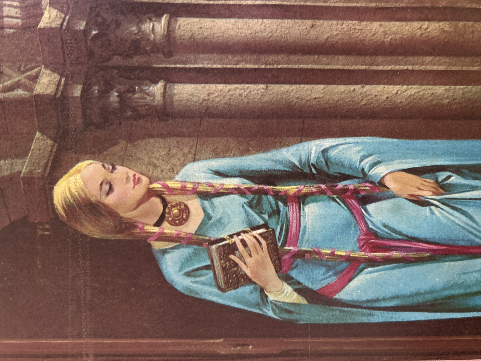 The image is a painting depicting a young woman with blonde hair braided with pink and gold ribbons. She is wearing a blue robe with a pink sash and a large, circular pendant necklace. In her left hand, she holds a closed, ornate book. The background appears to be an interior with architectural elements like columns and textured walls, suggesting a historical or fantastical setting. There is no identifiable information within the image or its context to suggest a location in Santa Ana, Venezuela.