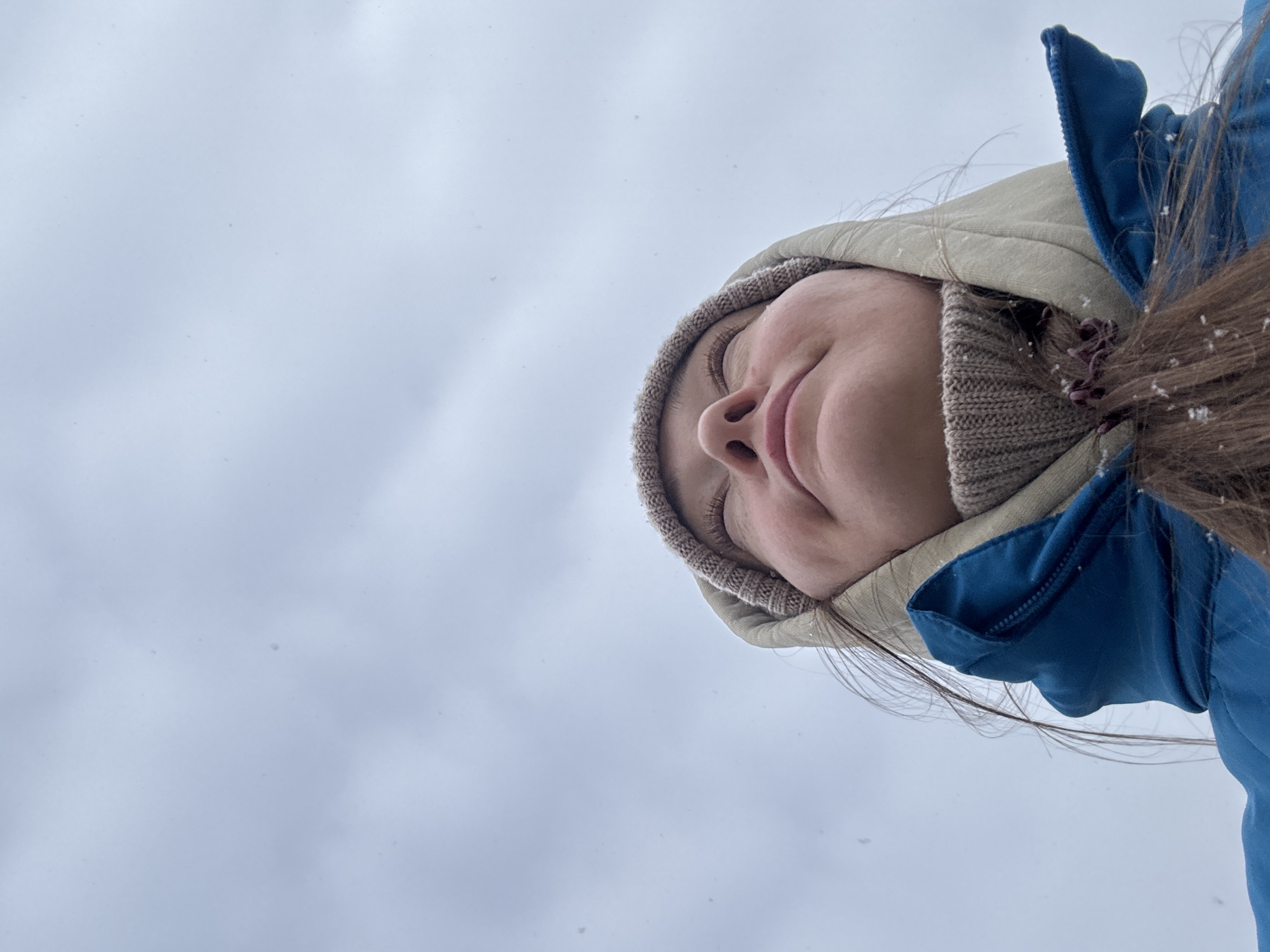 A low-angle shot shows a woman with closed eyes, wearing a light tan knit hat, a light tan hood, and a blue jacket. Snowflakes are visible on her hair. The background is a light gray, overcast sky. The location, Chaikovsky, Russia, cannot be verified from the image alone.