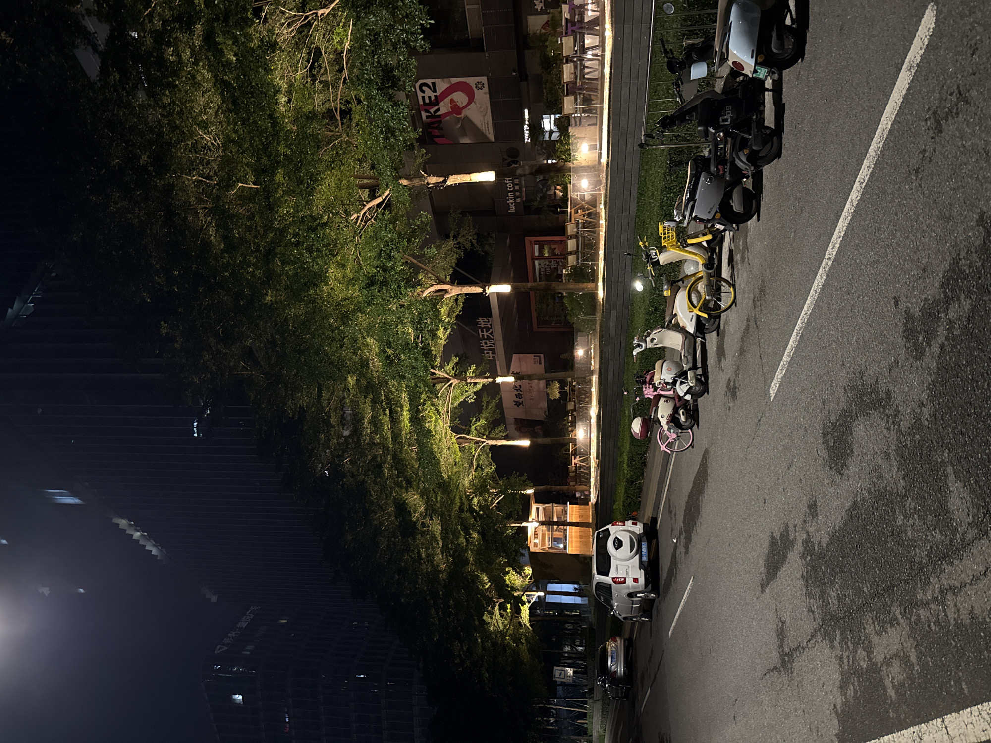 This is a night-time, outdoor photograph taken in the Haizhu District of China. The image captures a street scene with several parked electric scooters and a white SUV. Trees with dense foliage line the street, illuminated by streetlights and lights from nearby buildings. The ground appears to be asphalt, with a white line marking a parking space.
The scooters are of various colors, including white, yellow, and black, and some are decorated with pink accents. One scooter has a yellow basket on the front. The white SUV is parked on the right side of the image, facing away from the camera. Its license plate is visible, though the characters are not clearly discernible.
In the background, several buildings are visible, some with illuminated signs. One sign reads "luckin coffee" in English and Chinese. Another sign, partially obscured by trees, appears to be an advertisement with a large red graphic and text. The overall impression is one of a lively urban area at night, with businesses and transportation options readily available. The lighting suggests it is evening or night. There are no people visible in the frame.